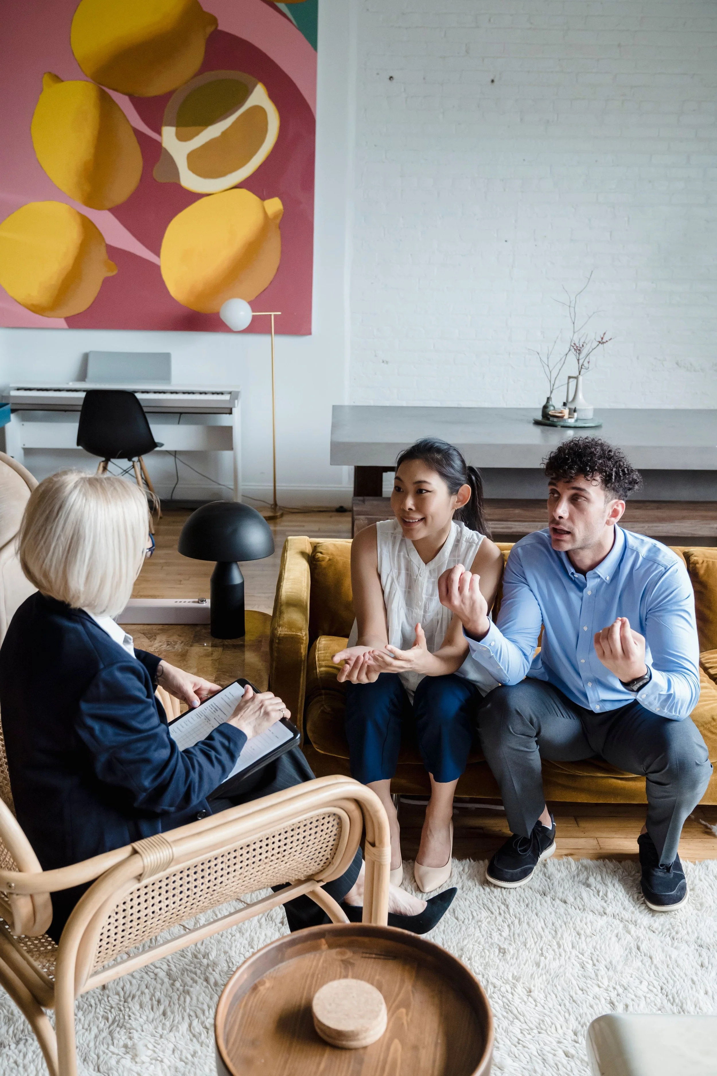 A group of four people sitting in a living room having a conversation. One woman is taking notes, another woman and man are talking animatedly, and a third woman is listening. The room has a white brick wall, a painting of lemons, a digital piano, a modern floor lamp, and a white shag rug.