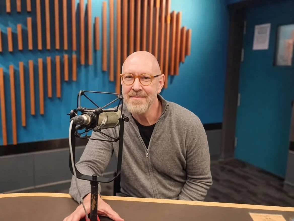 A man with glasses and a gray zip-up sweater sitting in front of a microphone inside a recording studio with blue walls and wooden acoustic panels.