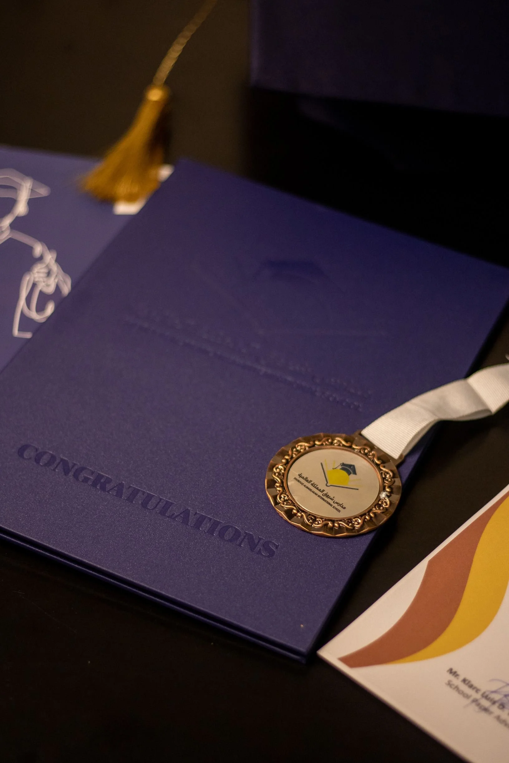A purple diploma or certificate with the word 'CONGRATULATIONS' embossed on it, a gold medal with Arabic text and a graduation cap logo, and a brochure or flyer with a yellow and brown color scheme.