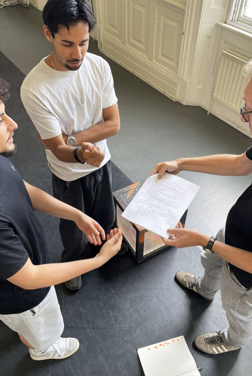 The photo is from above and shows two young men, on the left, looking at an older man, on the right, who is holding a script in his hand. It is from the rehearsal room. Two actors and a director.