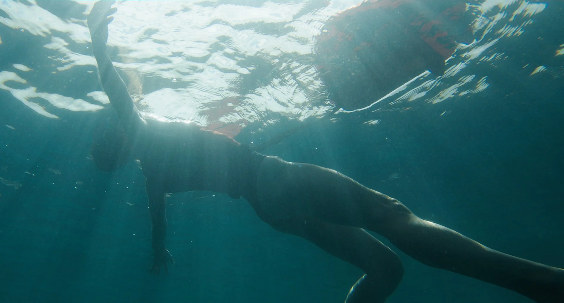 seeing a woman from underneath laying on her back floating in blue water, with the light shining through from above.