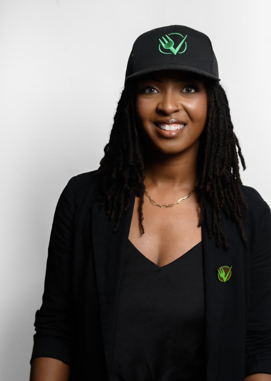 A smiling woman with dark curly hair wearing a black cap with a green logo, a black blazer with a matching logo pin, and a gold chain necklace against a plain white background.