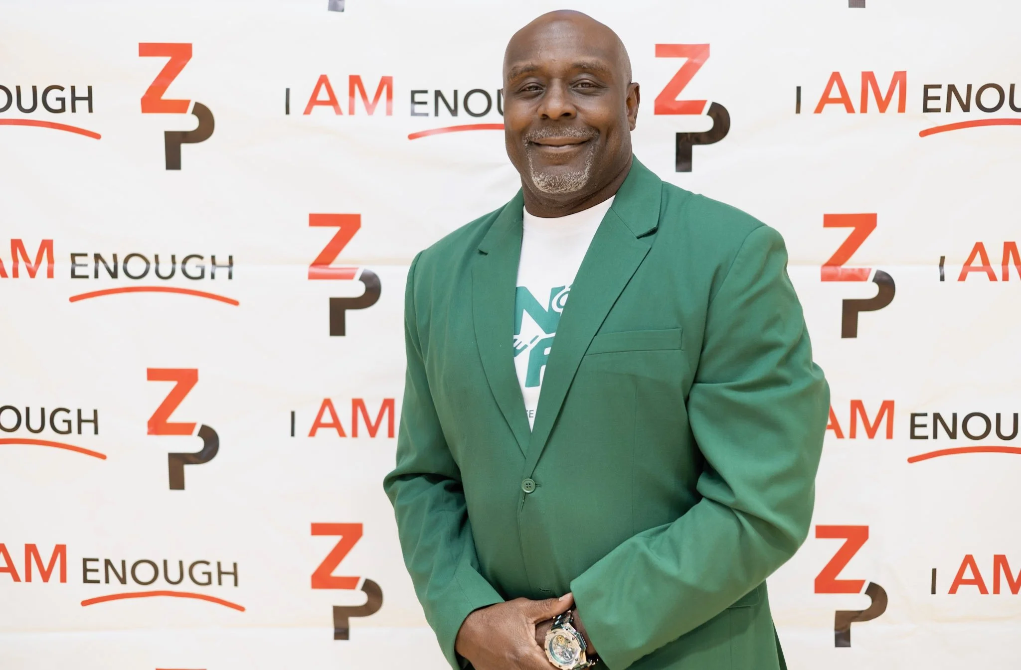A man in a green blazer standing in front of a backdrop with the words 'I AM ENOUGH' and the number 3 repeated. The man is smiling and has a wristwatch.