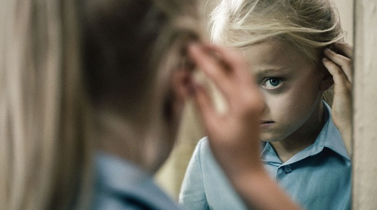 A young girl with blonde hair and blue eyes looking into a mirror, with her reflection seen clearly.