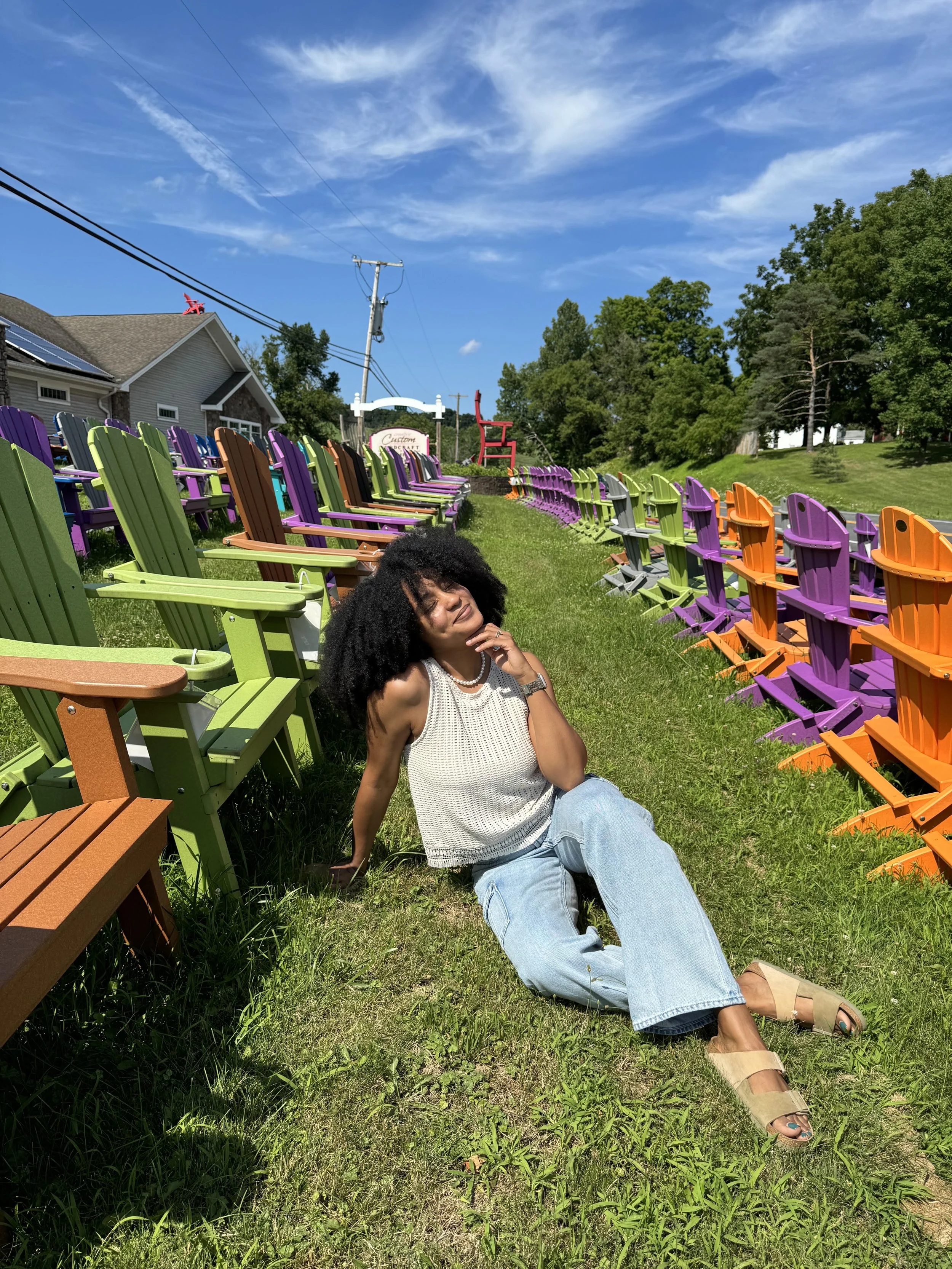 A smiling woman with curly hair sitting on the grass surrounded by colorful Adirondack chairs at an outdoor event under a blue sky with clouds.