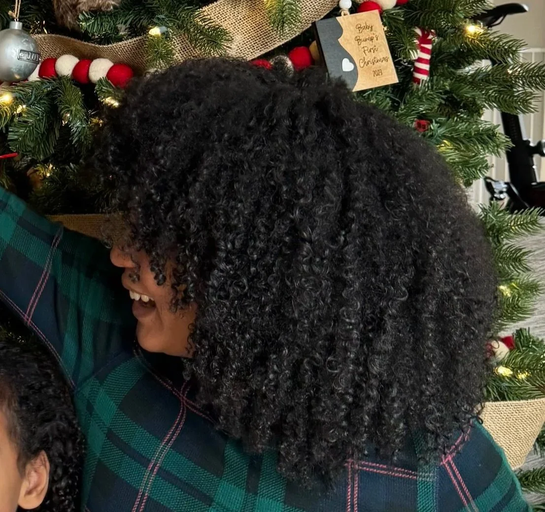 A family is celebrating Christmas in a living room. There is a large decorated Christmas tree with ornaments and lights. Some family members are sitting on the floor, others are holding children, and a woman is reaching up to hang a Christmas stocking on the wall. The scene is festive and joyful.