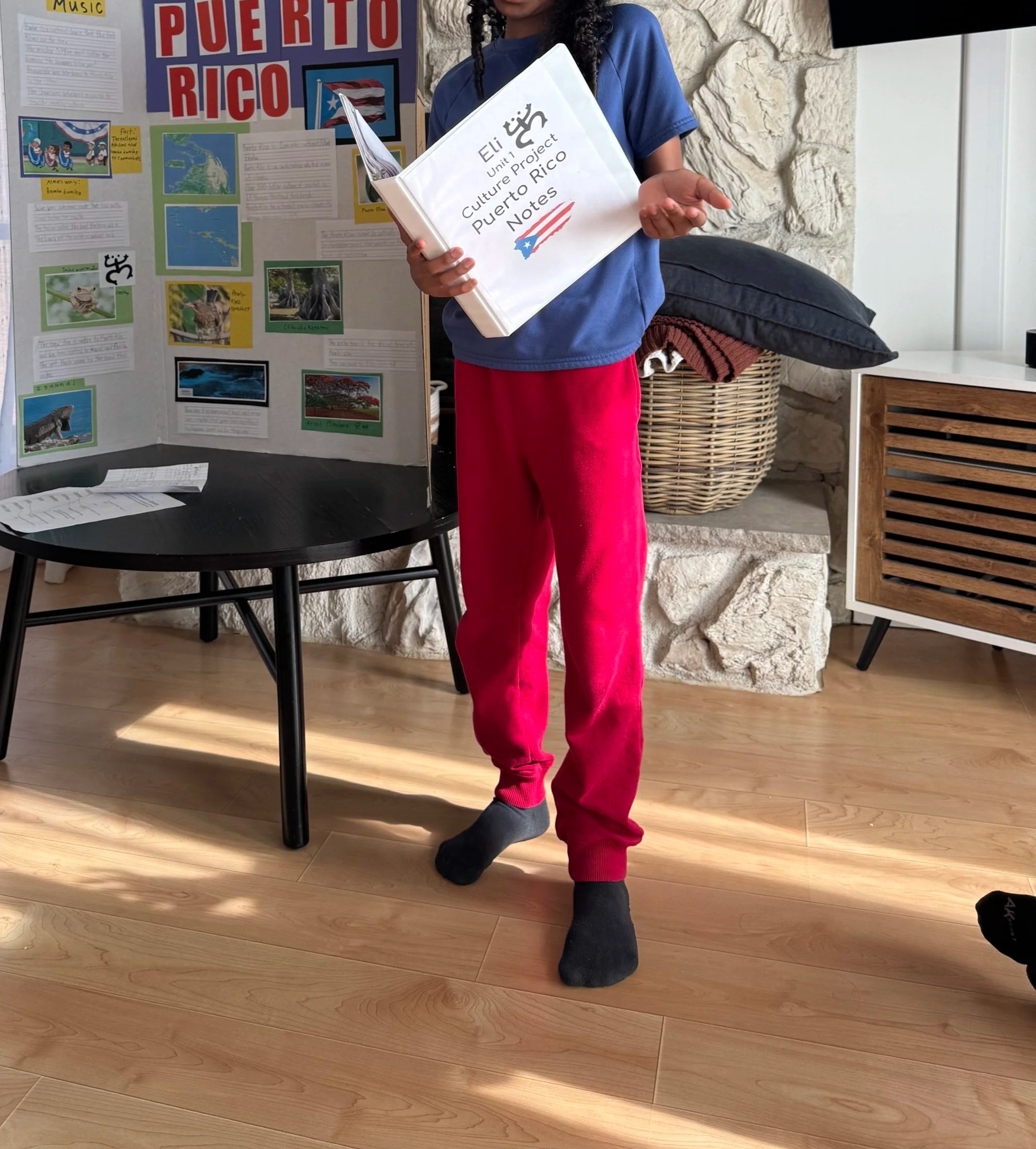 A young person stands in a living room next to a display board with information about Puerto Rico. They are holding a notebook titled 'Eli Unit 4 Culture Project Puerto Rico Notes' and wearing a colorful bucket hat, a blue shirt, and bright red sweatpants. They are barefoot on a wooden floor with sunlight casting shadows in the room.
