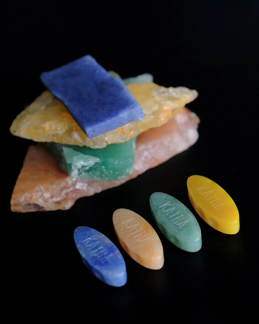 A collection of aventurine stones stacked on top of each other with laser-cut aventurine beads in corresponding colors showing "KATRA" engraving on the back side of the beads on a black background.