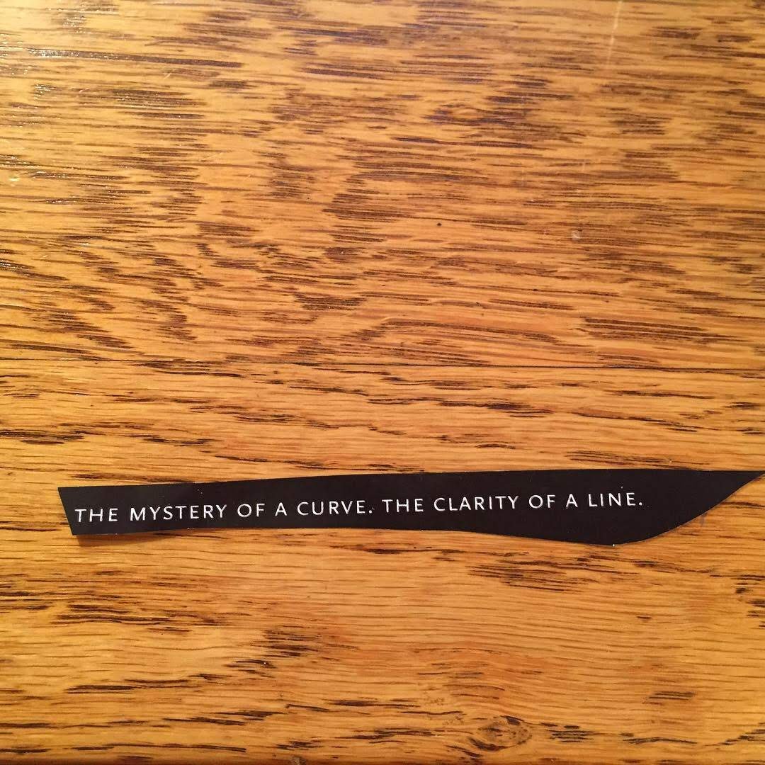 A black paper strip with white text that reads, "The mystery of a curve. The clarity of a line," placed on a wooden surface.
