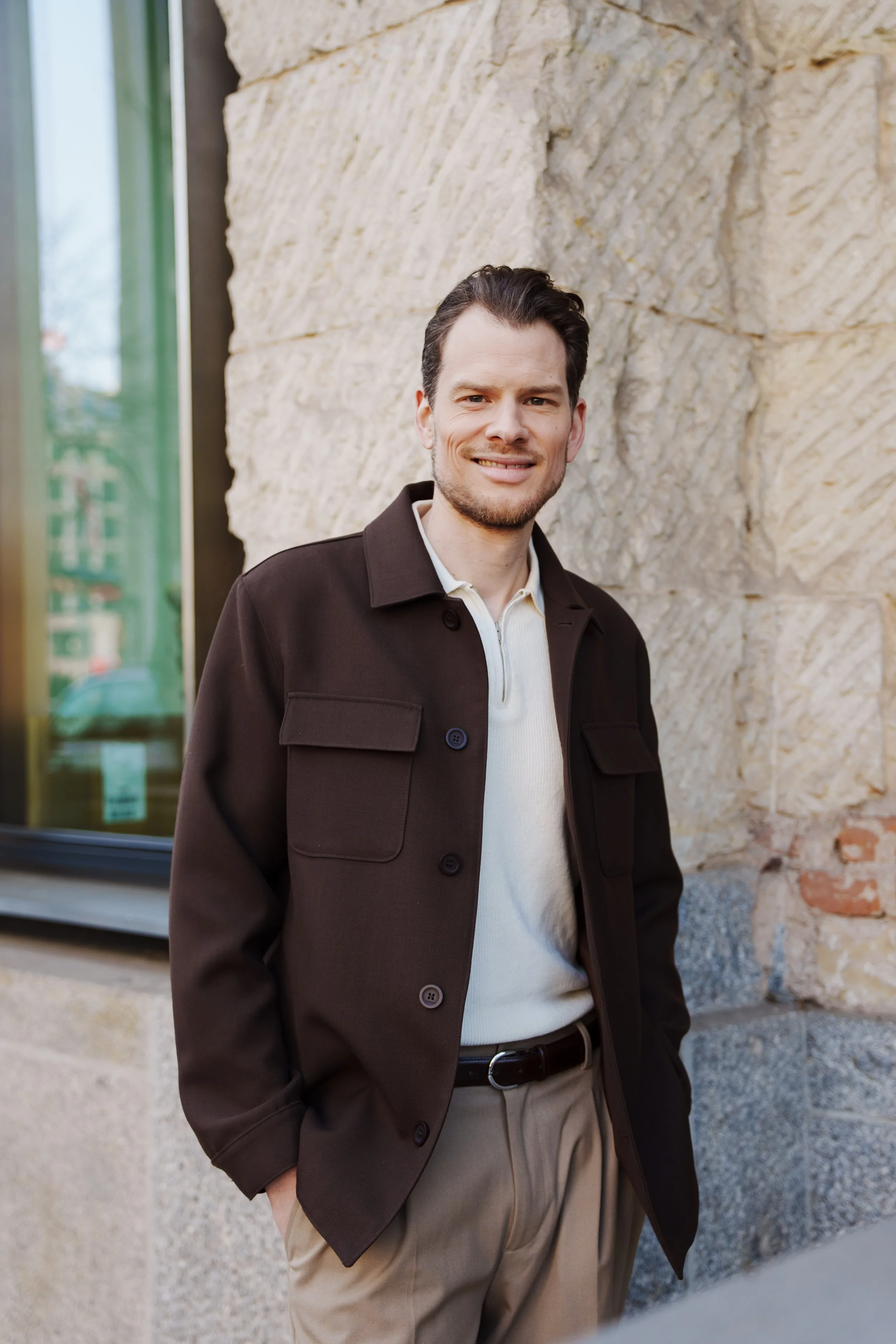 Ein Mann steht vor einer Steinmauer und lächelt, trägt eine braune Jacke, ein weiss- cremefarbenes Shirt und beige Hosen.