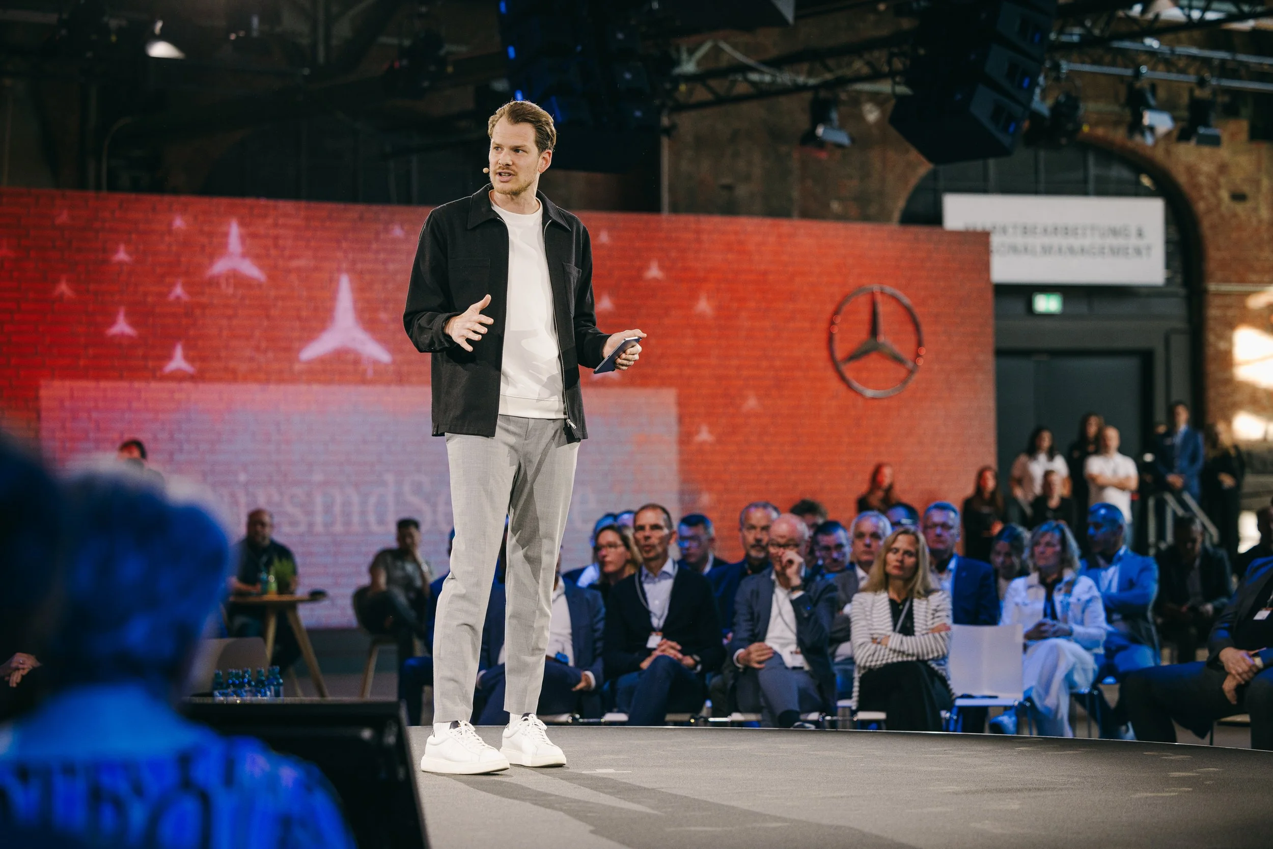 Mann hält Vortrag vor Publikum auf einer Bühne in einem großen, industrialähnlichen Veranstaltungsraum mit rotem Backsteinmauer im Hintergrund und Mercedes-Logos, während die Zuhörer aufmerksam zuhören.