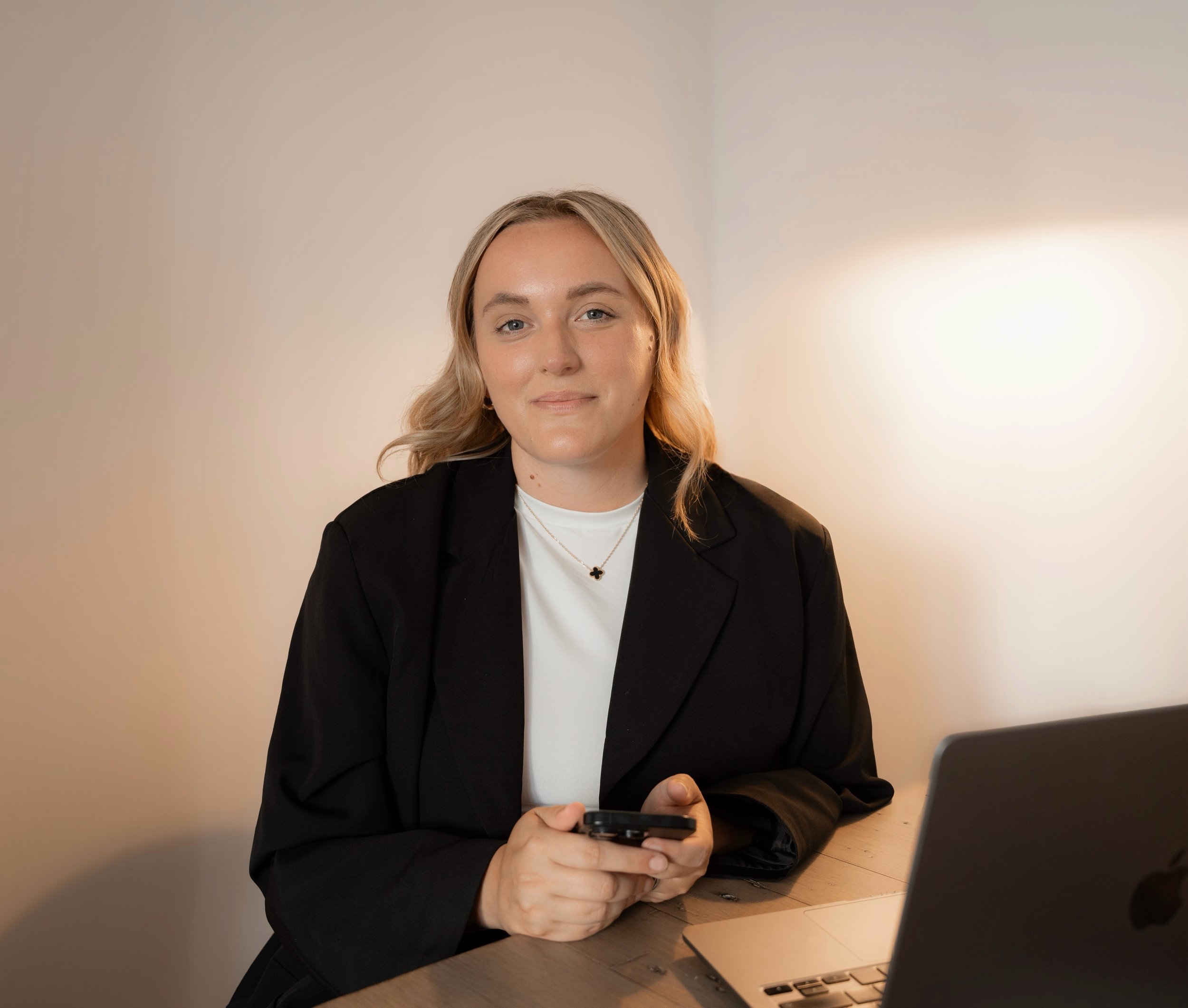 A young woman with blonde hair, wearing a black blazer and white shirt, seated at a wooden table, holding a smartphone, with a laptop partially visible on the table in front of her, and a neutral-colored wall in the background.
