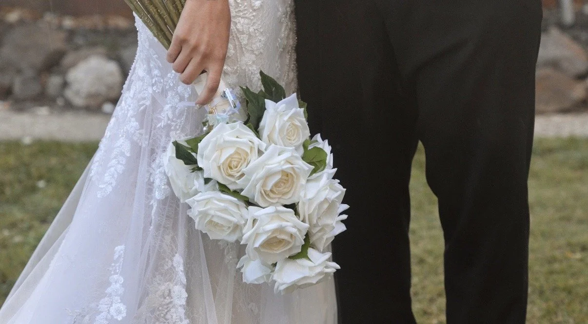 Bride holding a bouquet of white roses, standing next to groom in black suit outdoors.