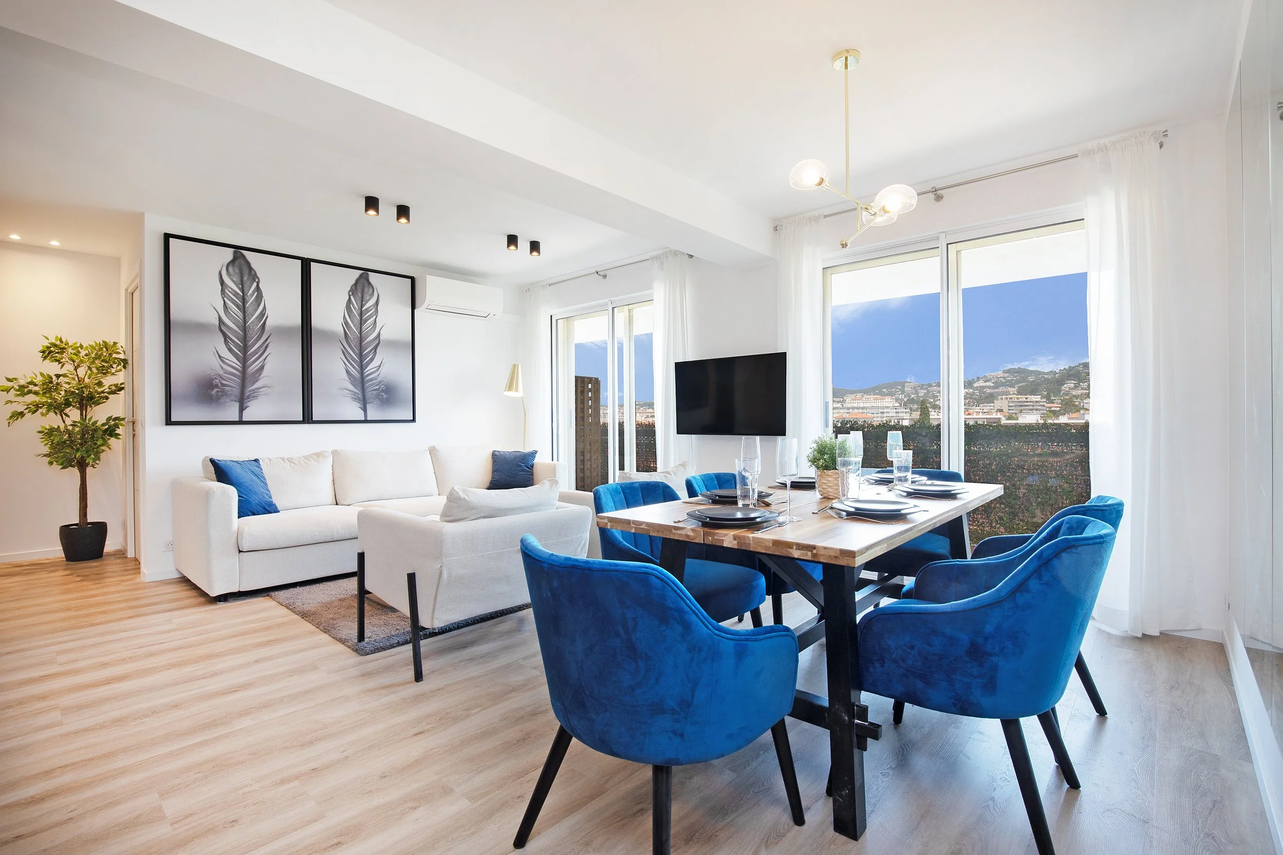 Modern living and dining room with a white sofa, blue chairs, wooden table, and large feather artwork on wall. Bright space with large windows and mountain view.