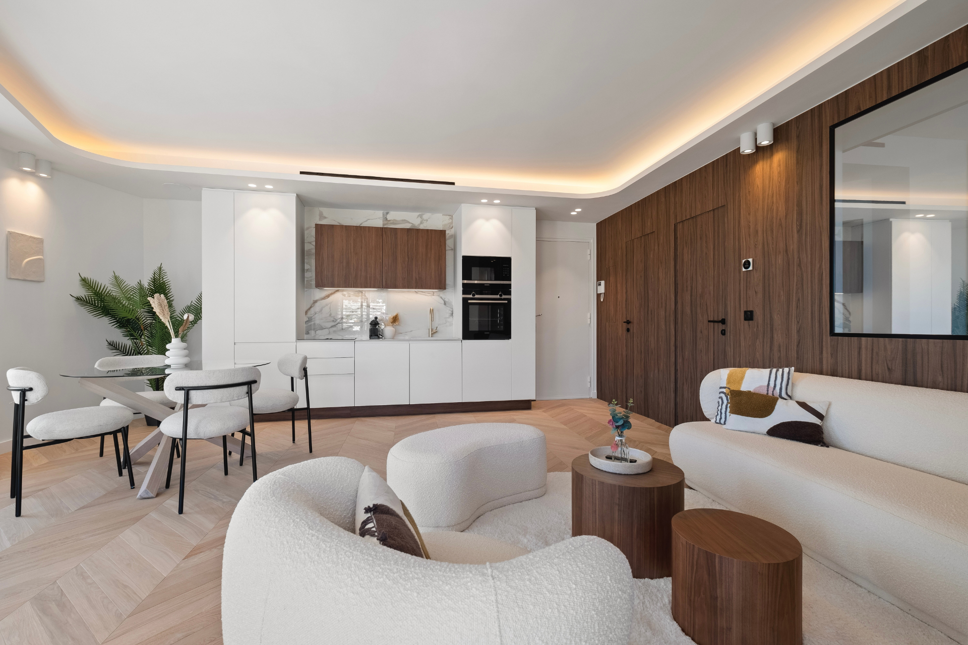 Modern living room with open-concept design, featuring a kitchen area with white and wooden cabinets, a dining table with four chairs, and a lounge area with a white sofa and armchairs. A plant decorates the corner, and the room has warm lighting and wood flooring.