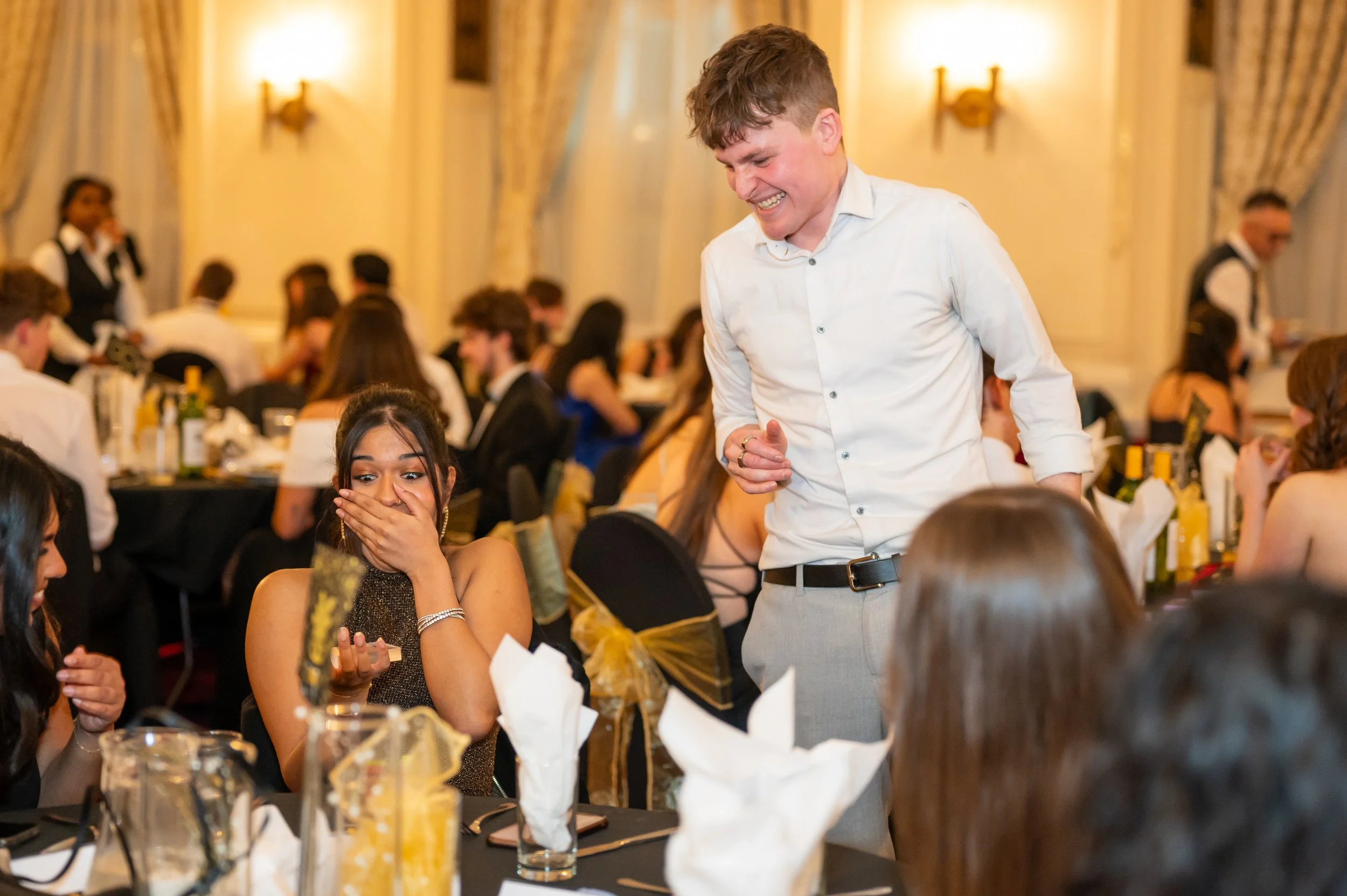 People laughing and reacting at a formal event with elegantly dressed tables in the background.