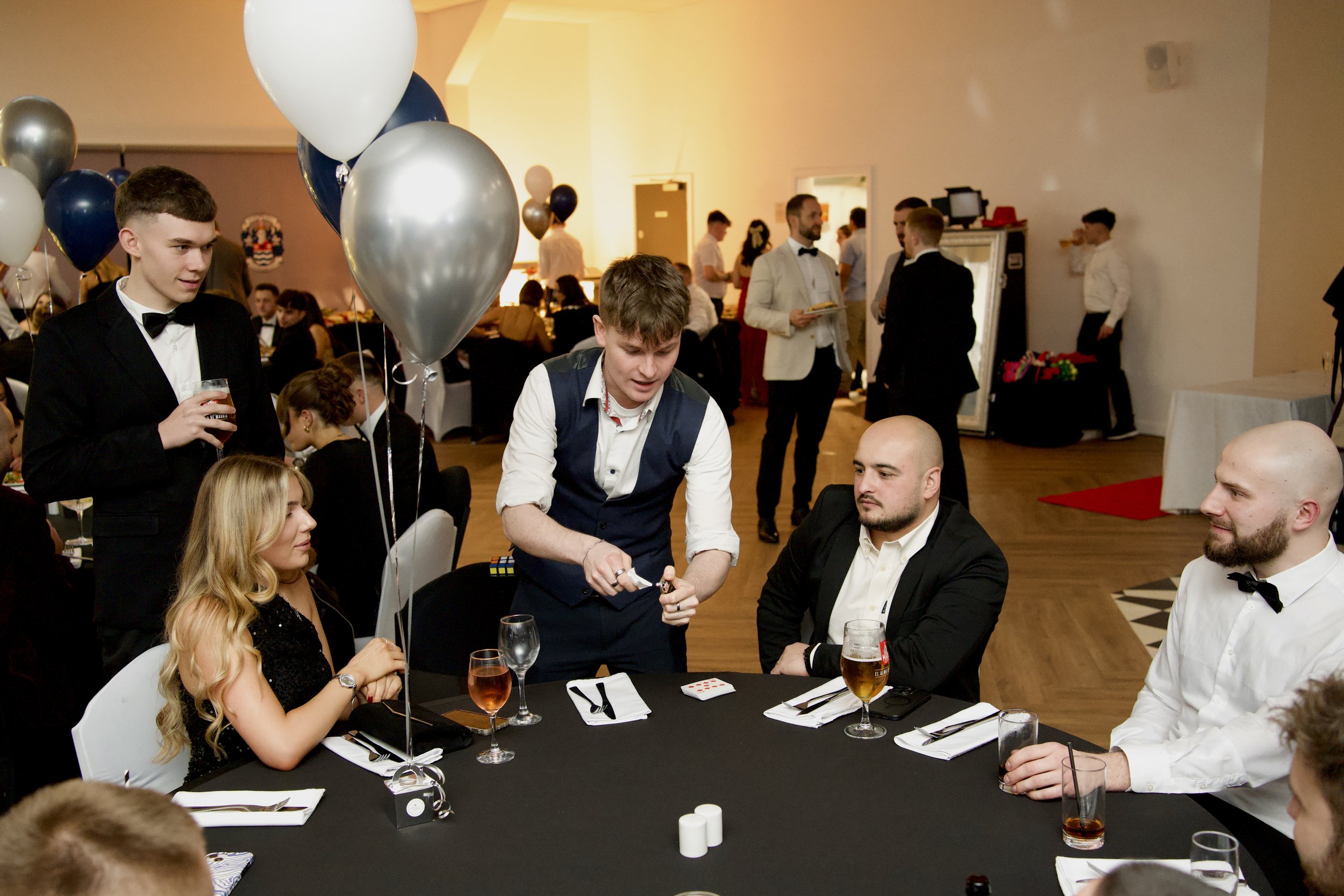 People in formal attire attending a magic performance at a banquet, seated around a table with drinks and silver balloons.