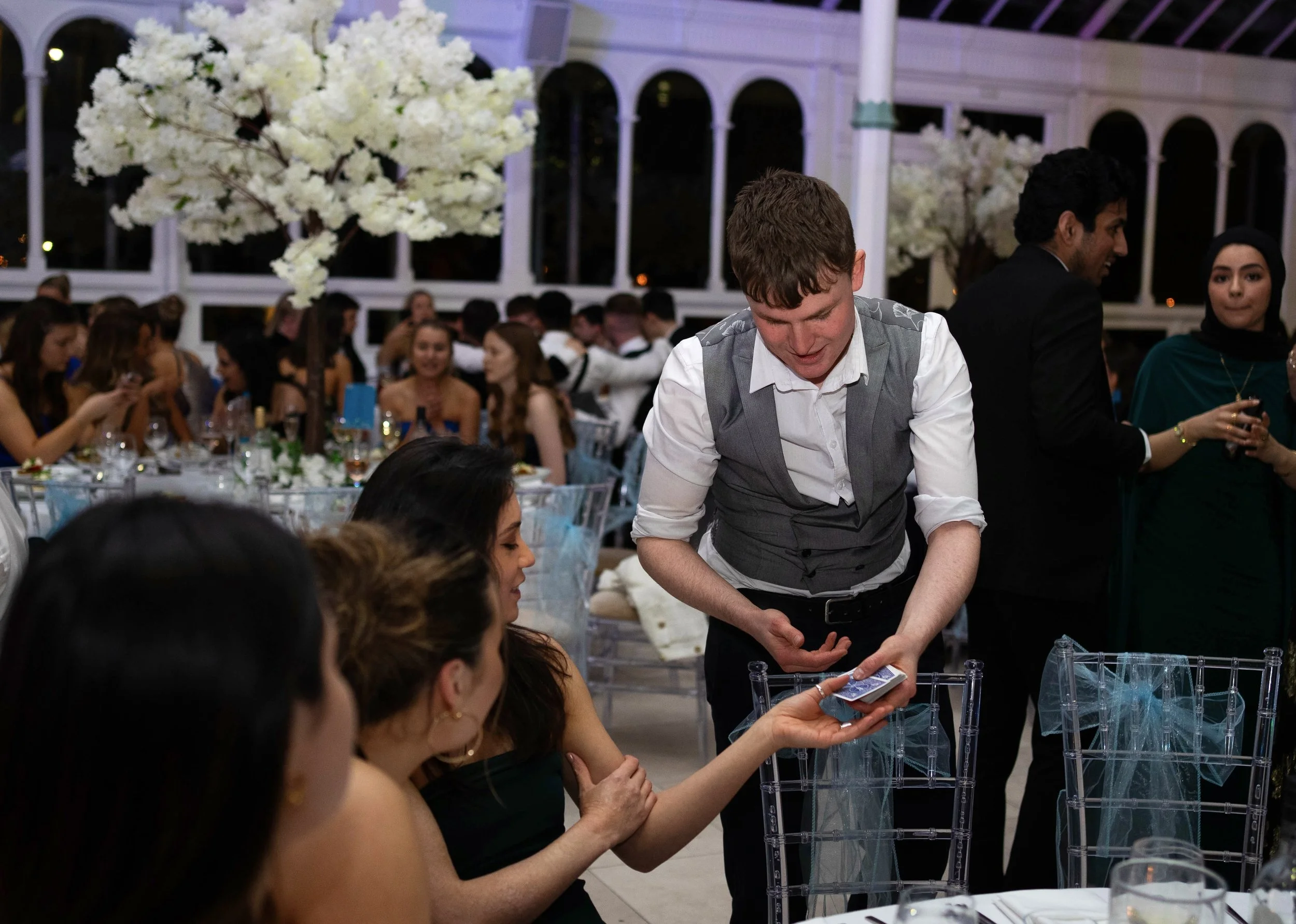 Daniel Maze performing a card trick for seated guests at a formal event, with a decorative floral centerpiece and elegant decor in the background in Liverpool.