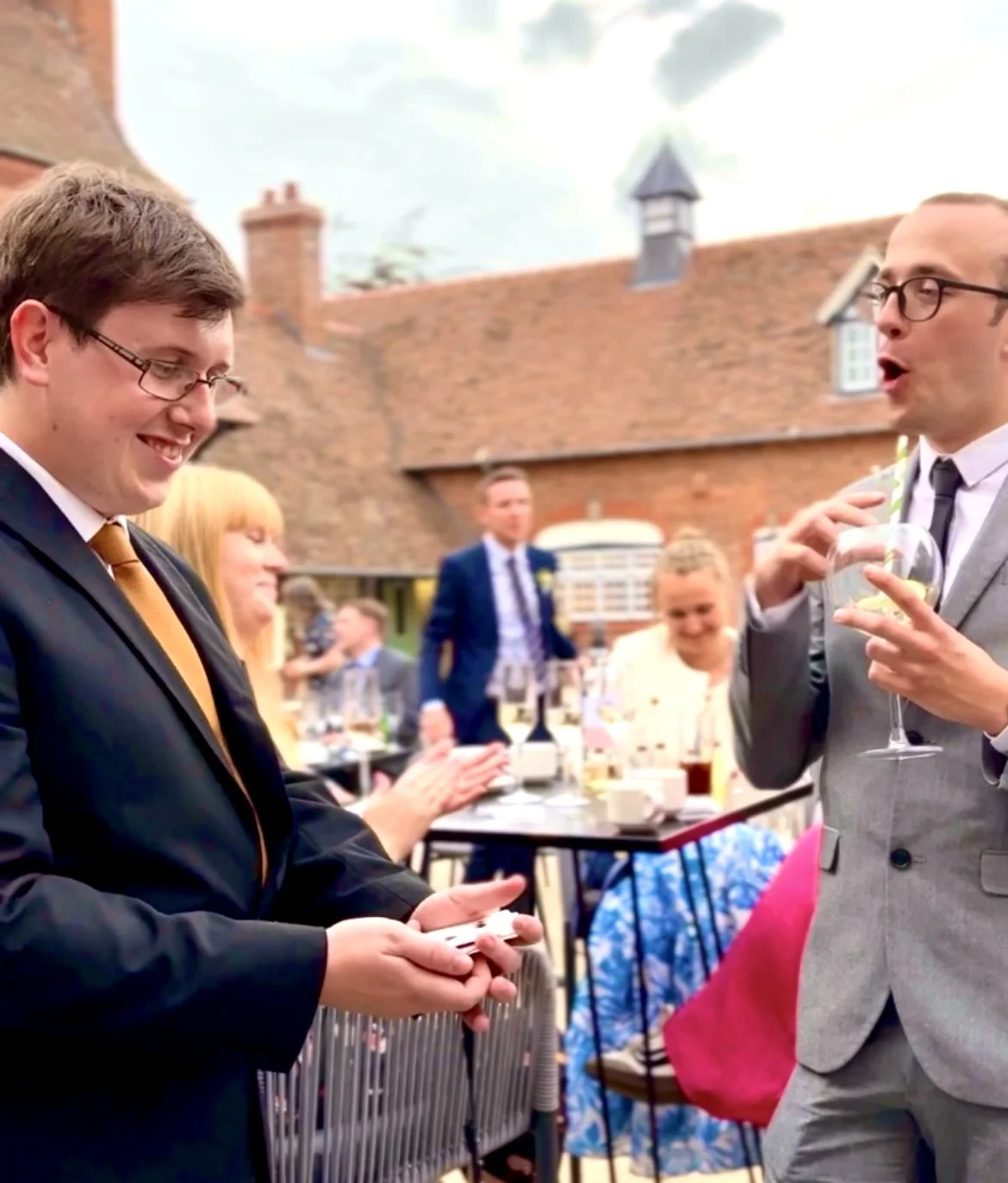 Outdoor Wedding Day with man in black suit performing card trick for attendees wearing formal attire. 