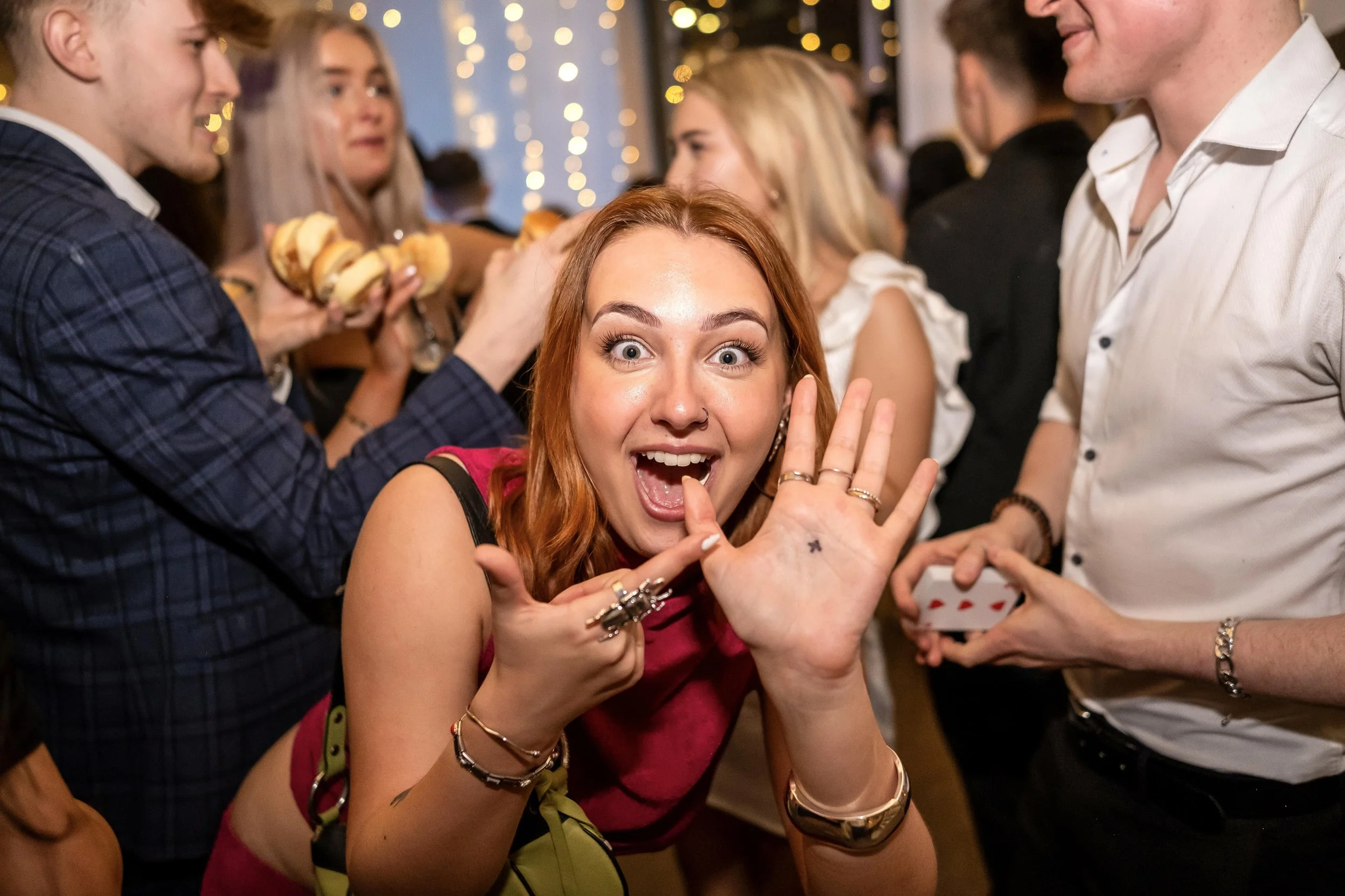 People having fun at a party with a woman in the foreground excitedly showing her palm, while others socialize in the background, one holding sliders and another with playing cards after an astounding magic trick.