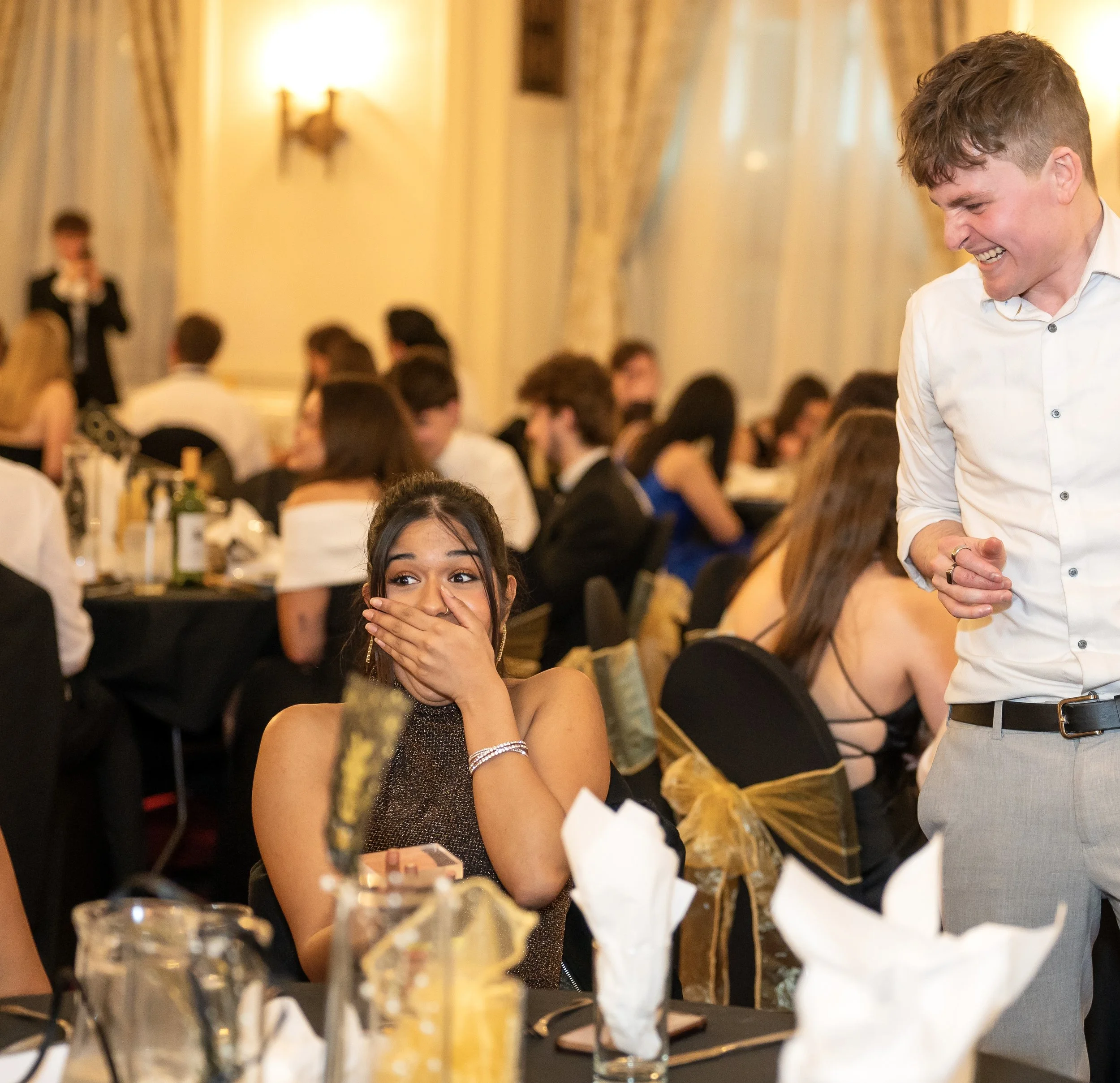 People at a formal event, a woman looking surprised, and a man smiling in a banquet setting with tables and decorations as Daniel performs unforgettable magical entertainment. 