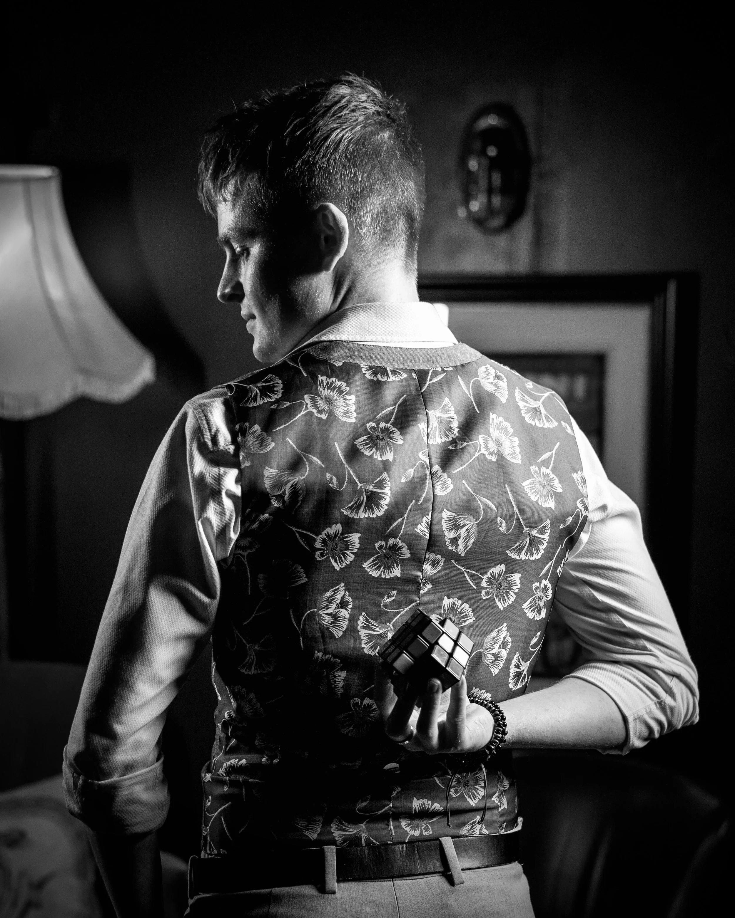 Black and white image Daniel Maze holding a Rubik's Cube behind his back, standing in a room with a lamp and framed picture. Taken at The Oracle In Liverpool.