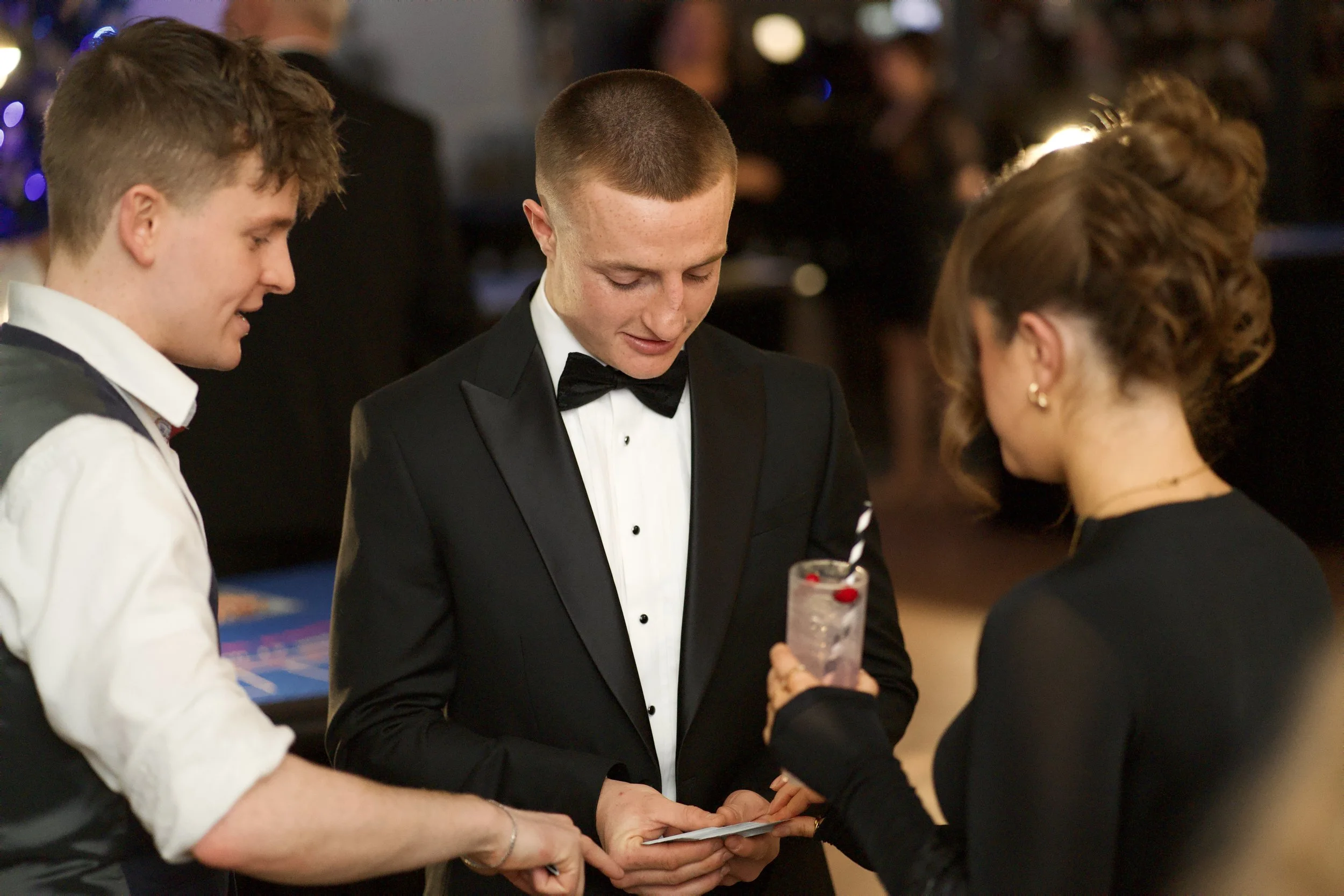 Three people at a formal event, with one man in a black tuxedo holding a card, another man in a white shirt and vest, and a woman watching Daniel Maze perform incredible magic. 