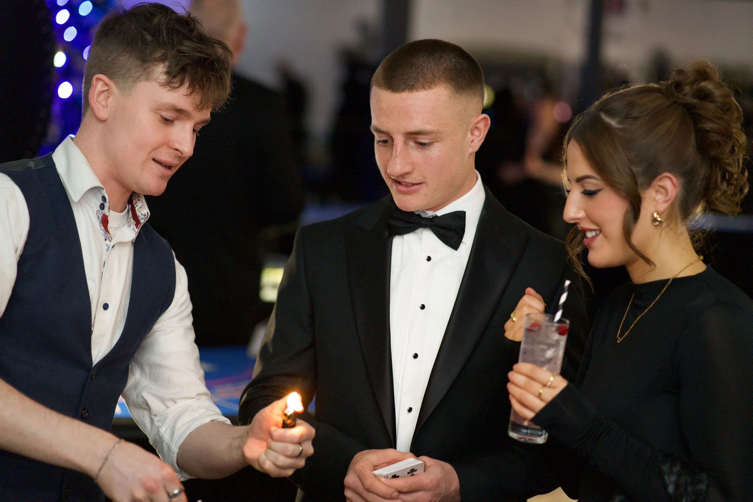 Three people in formal attire, one holding a deck of cards, another holding a drink, and the third using a lighter. Manchester Wedding Venue. 