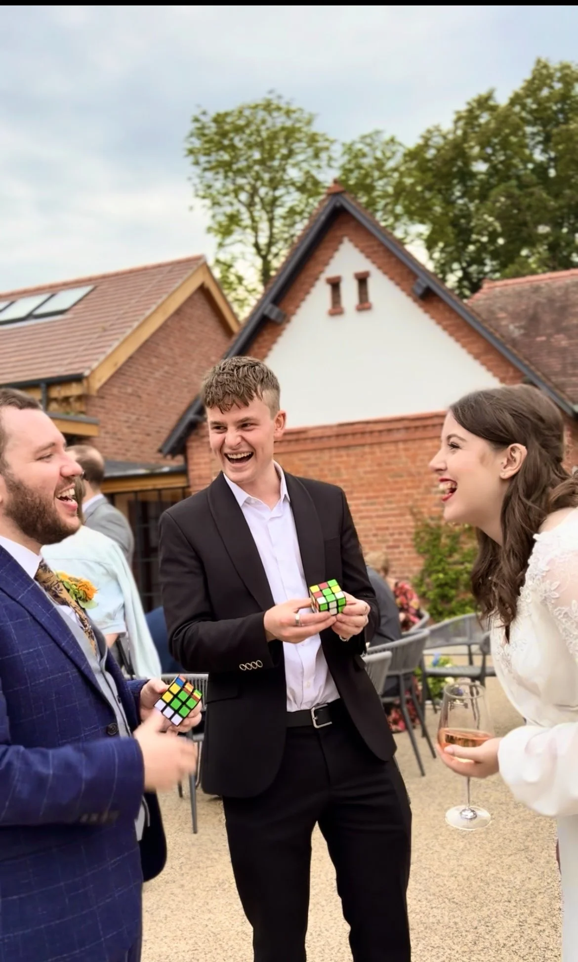 Wedding couple and magician outside laughing, two holding Rubik's Cubes, one with a wine glass; beautiful weather and formal attire. 