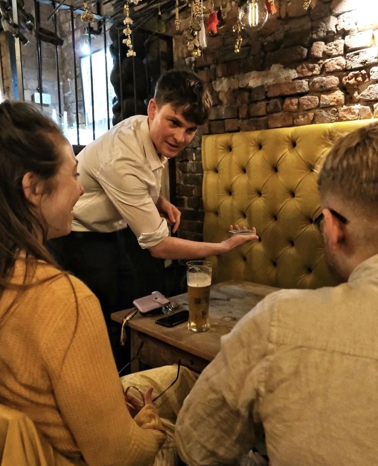 Two people in a cozy bar interact with a performer displaying a card trick, while seated at a rustic table with drinks under warm ambient lighting in Liverpool.