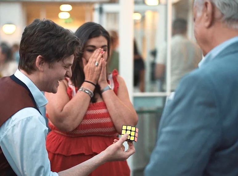Daniel Maze solving a Rubik's cube while two women watch and react with surprise and joy, in an indoor setting.