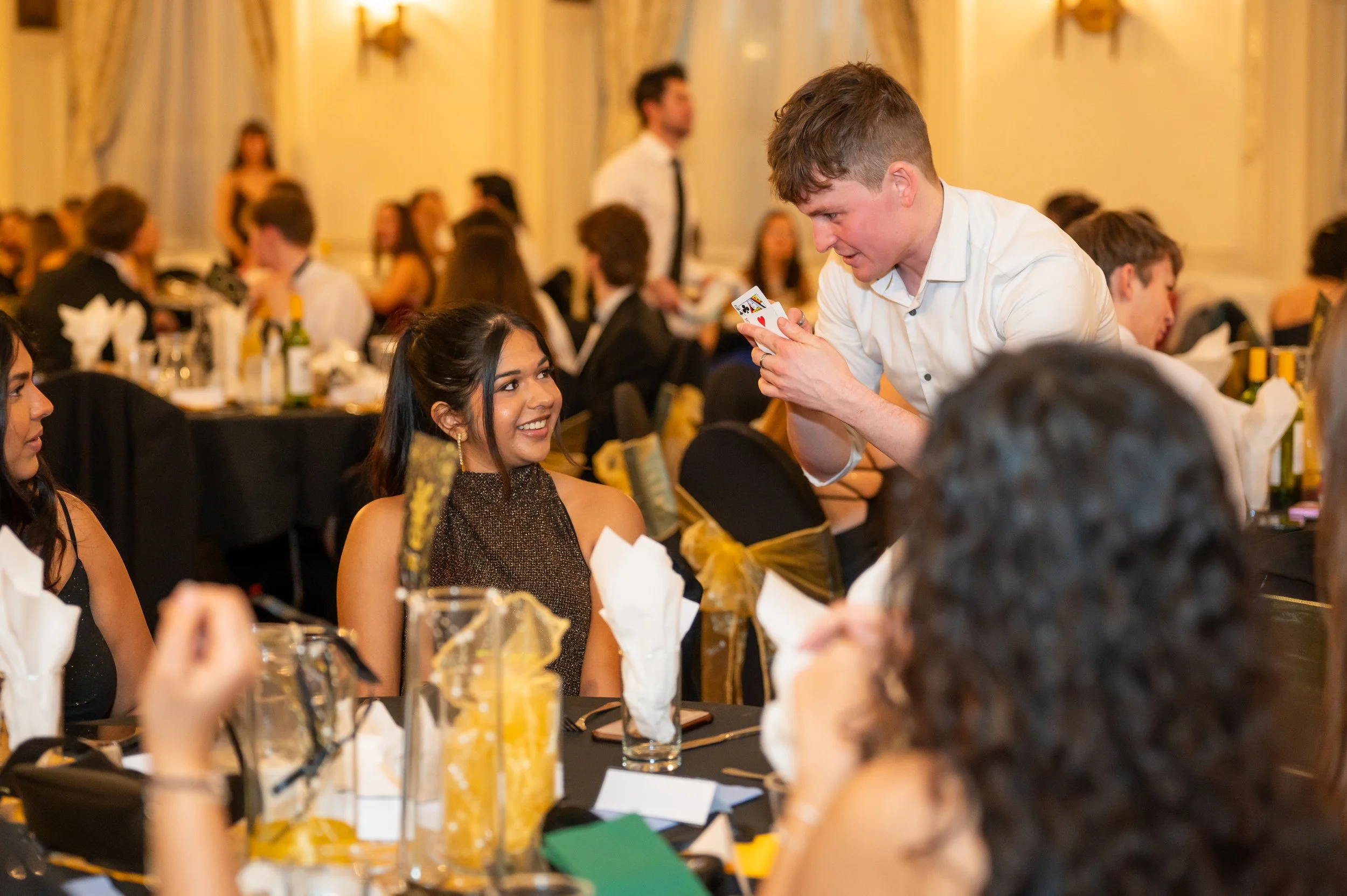 A magician performs a card trick at a formal event while a young woman watches, smiling. The setting is a banquet hall with elegantly decorated tables and attendees dressed in formal attire. Daniel Maze, the magic man of Manchester.