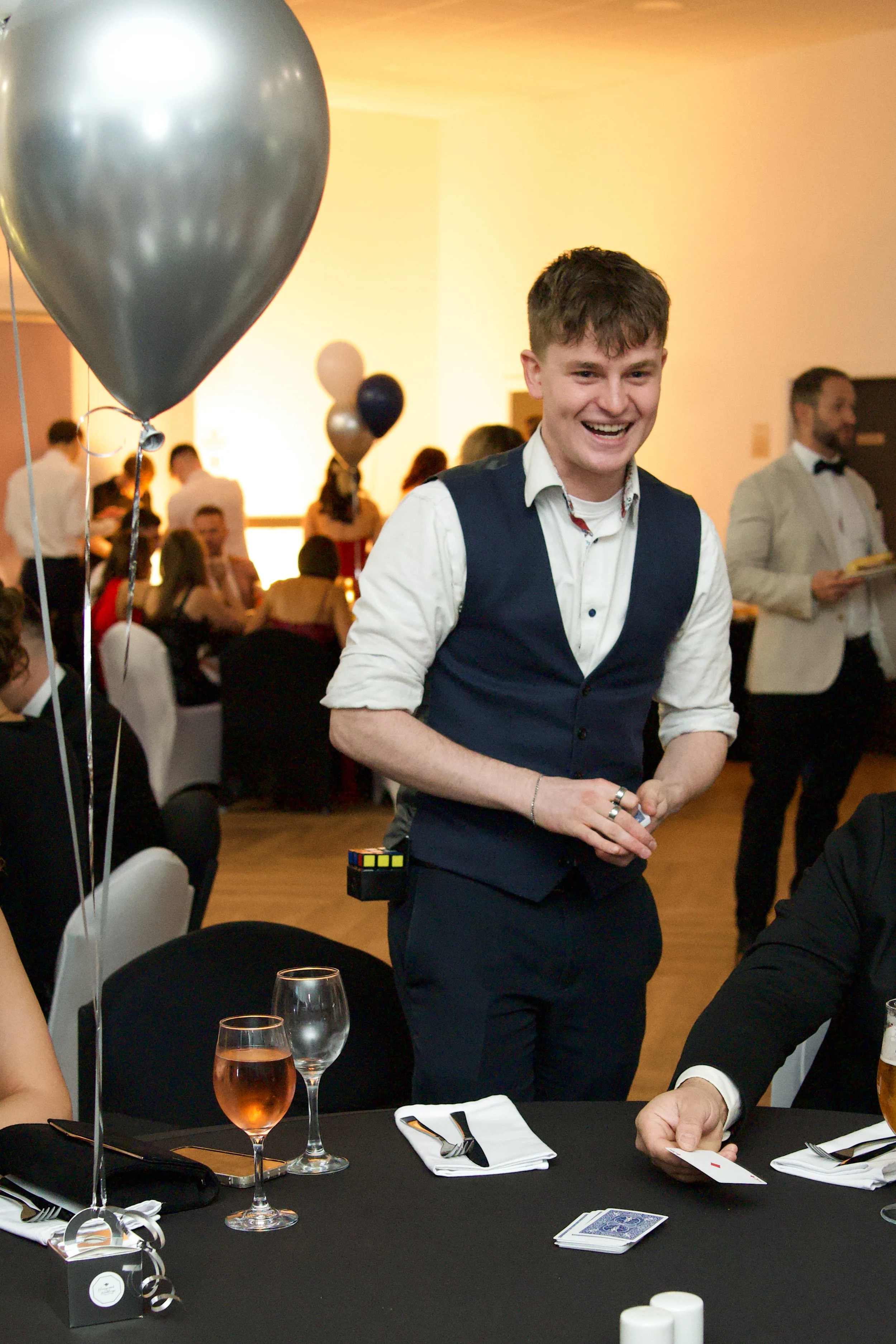 Wedding Magician Daniel Maze in formal attire performing a card trick at a party with balloons and people in the background.