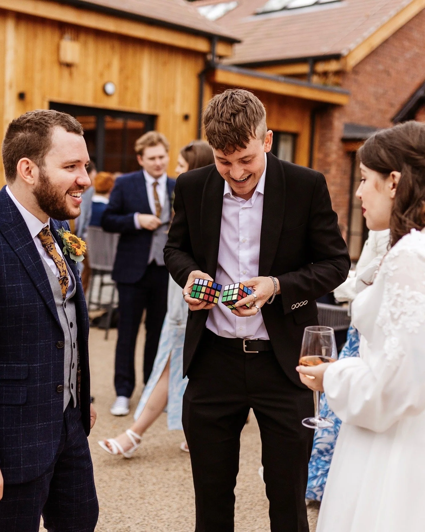 Group of people at an outdoor event, one holding two Rubik's cubes, another holding a wine glass. Everyone is dressed in formal attire. Background shows a wooden building.