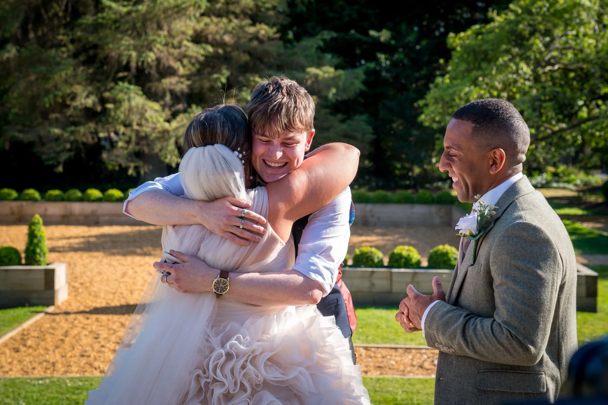 A couple getting married, smiling and hugging, while a man in a beige suit observes happily in a garden setting. Daniel Maze and bride hugging it out. 