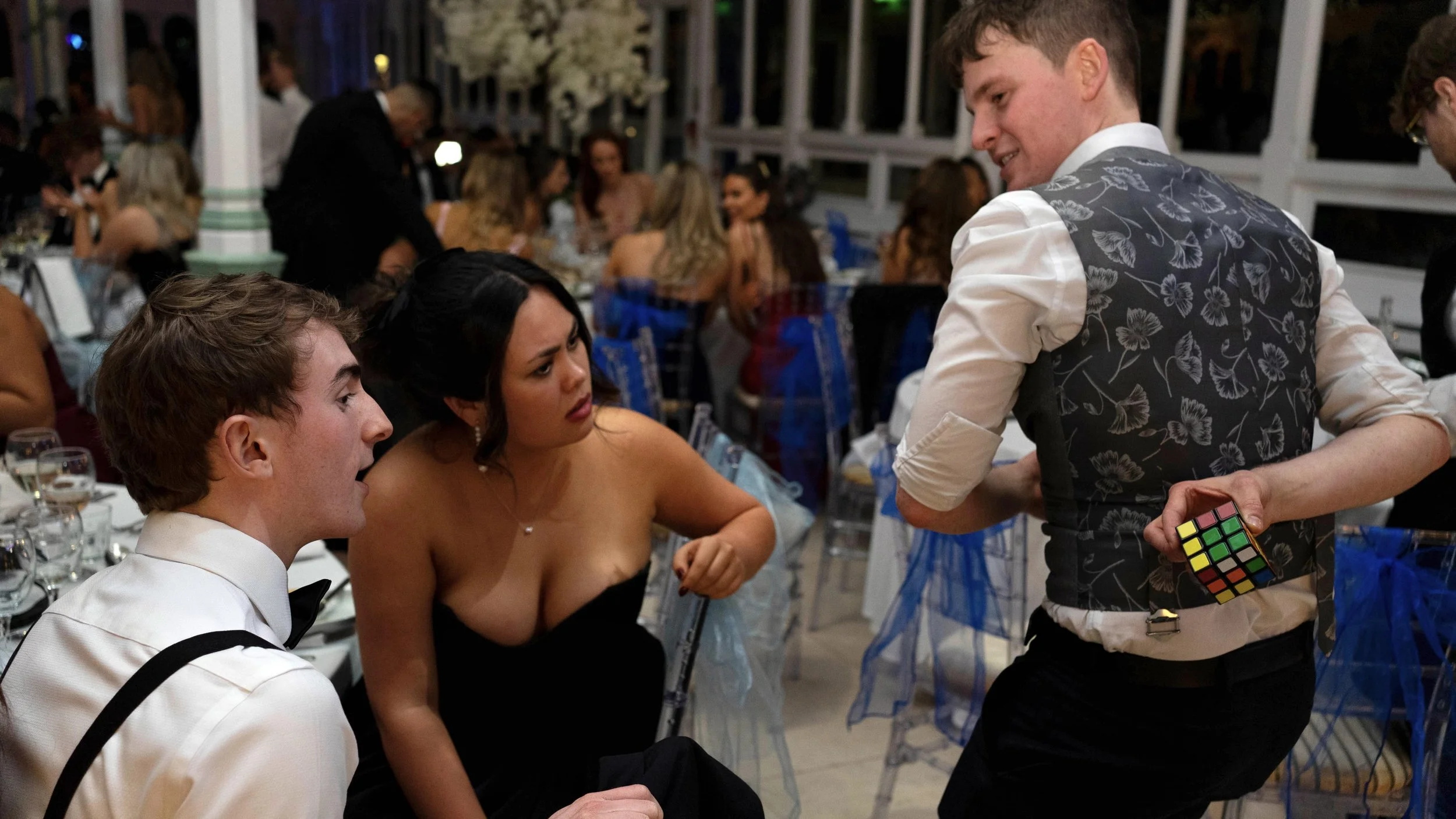 Daniel Maze magician holds a Rubik's Cube behind his back while two seated guests, a man and a woman, look on with interest at a formal event with many guests behind at the Wedding Day.