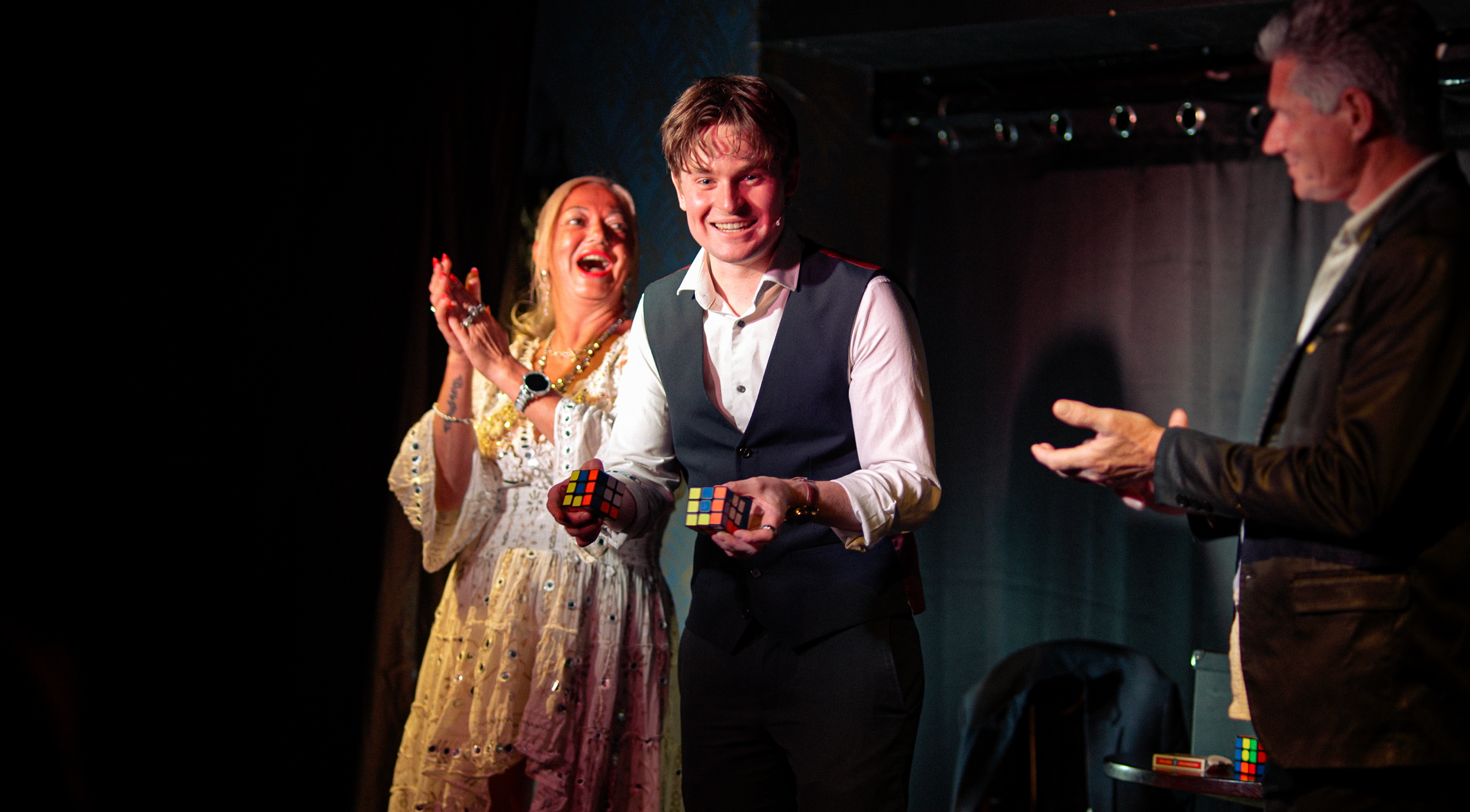 Daniel Maze smiling and holding two Rubik's cubes on stage, flanked by a woman clapping and an older man gesturing, during a lively event - The Unseen Room, Manchester. 