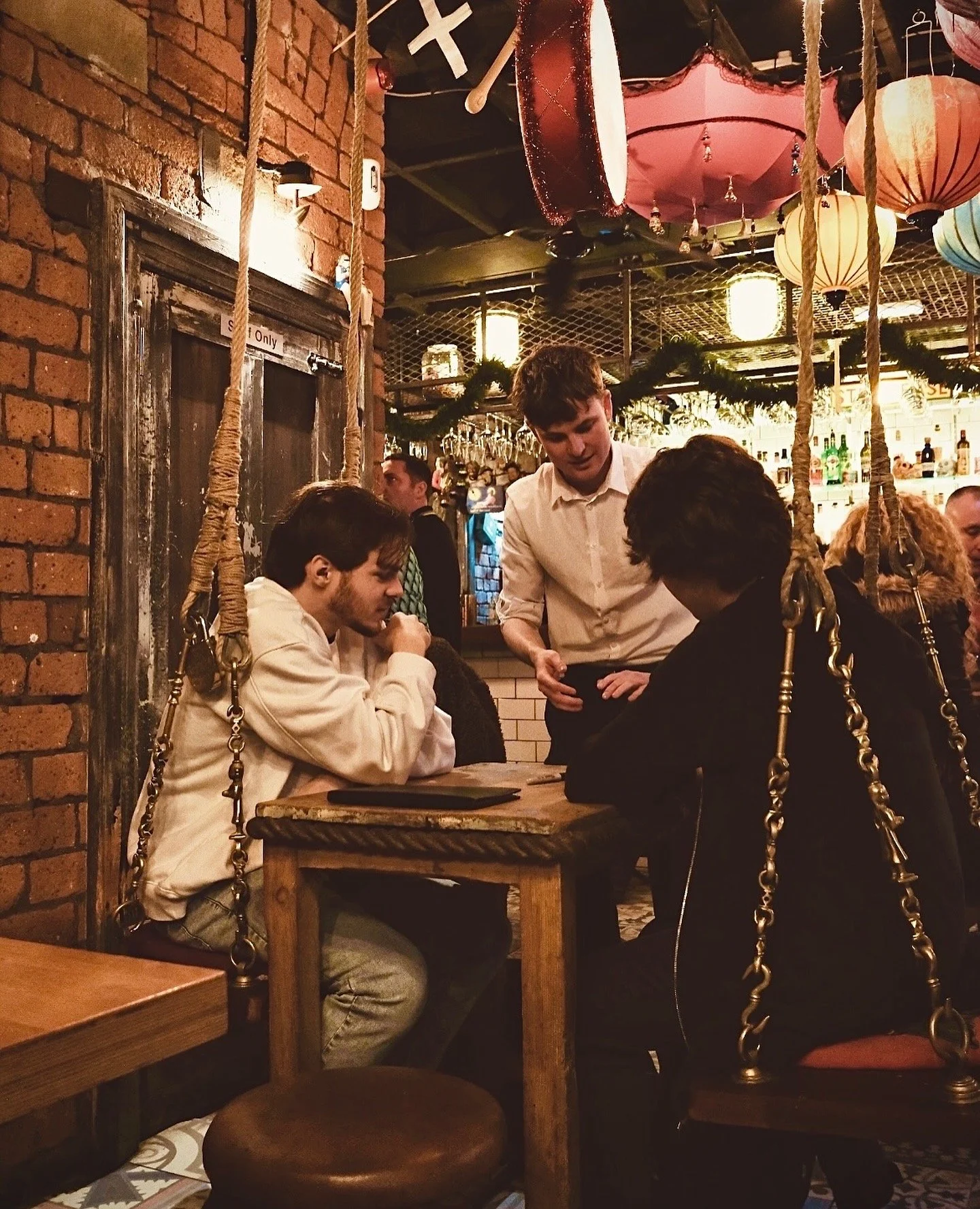 Cozy café with eclectic décor featuring people seated at a wooden table on swing seats, and a waiter taking orders. Vintage brick walls and colorful lanterns add to the ambiance in Liverpool.
