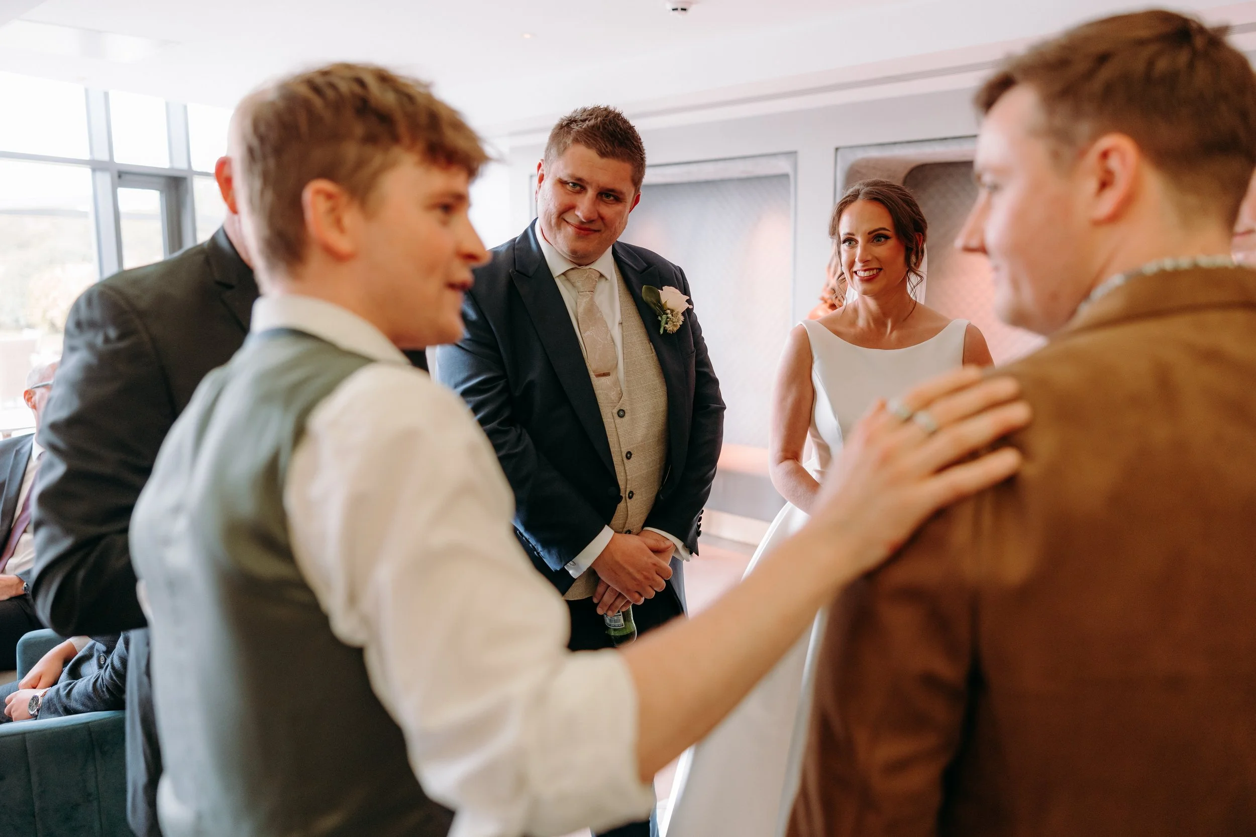 A group of people at a Wedding smiling and laughing including a bride in a white dress and a groom in a dark suit, standing indoors and conversing with Daniel Maze, magician. 