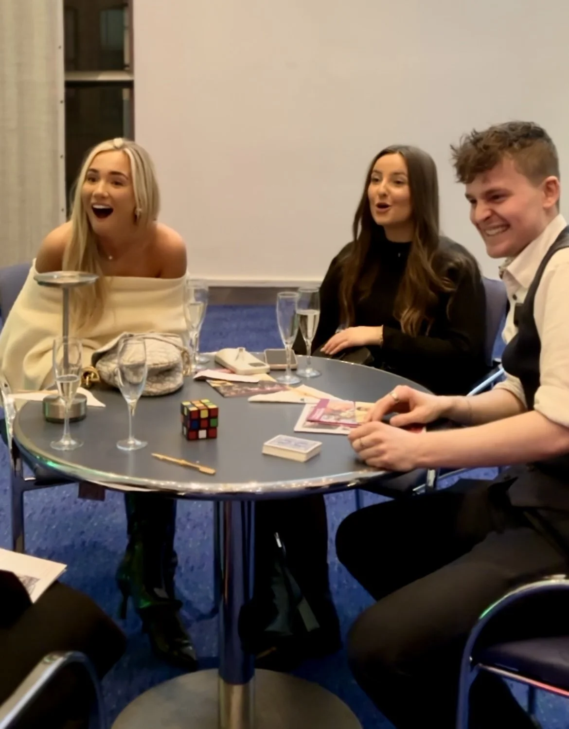 Group of three people seated at a table with drinks, a Rubik's cube, and playing cards.