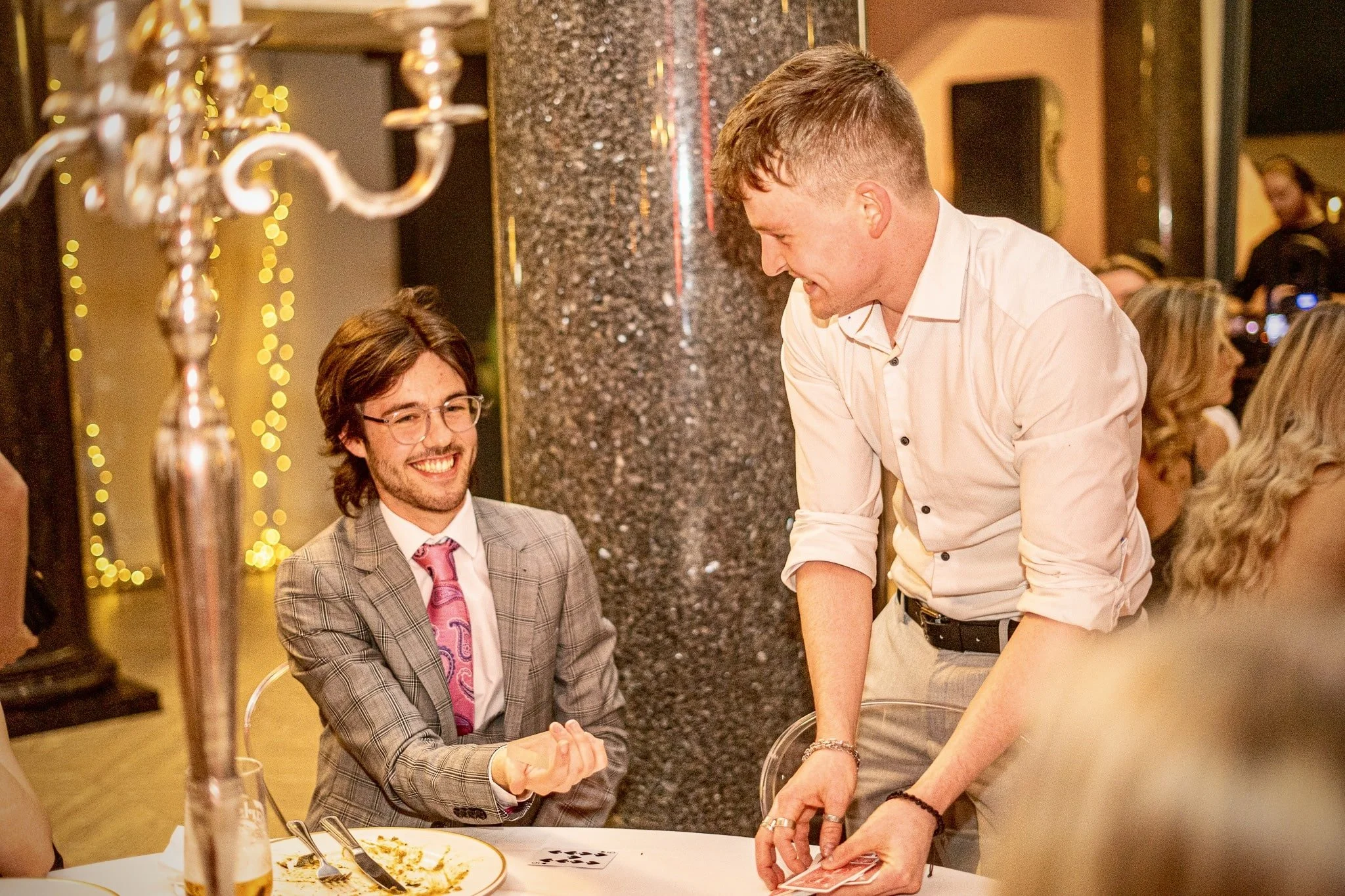 Two men at a table during a formal event, one sitting and smiling, wearing glasses, a checked suit, and a pink tie, the other standing and performing a card trick, wearing a white shirt, in a dimly lit elegant setting with a silver candelabra and blu