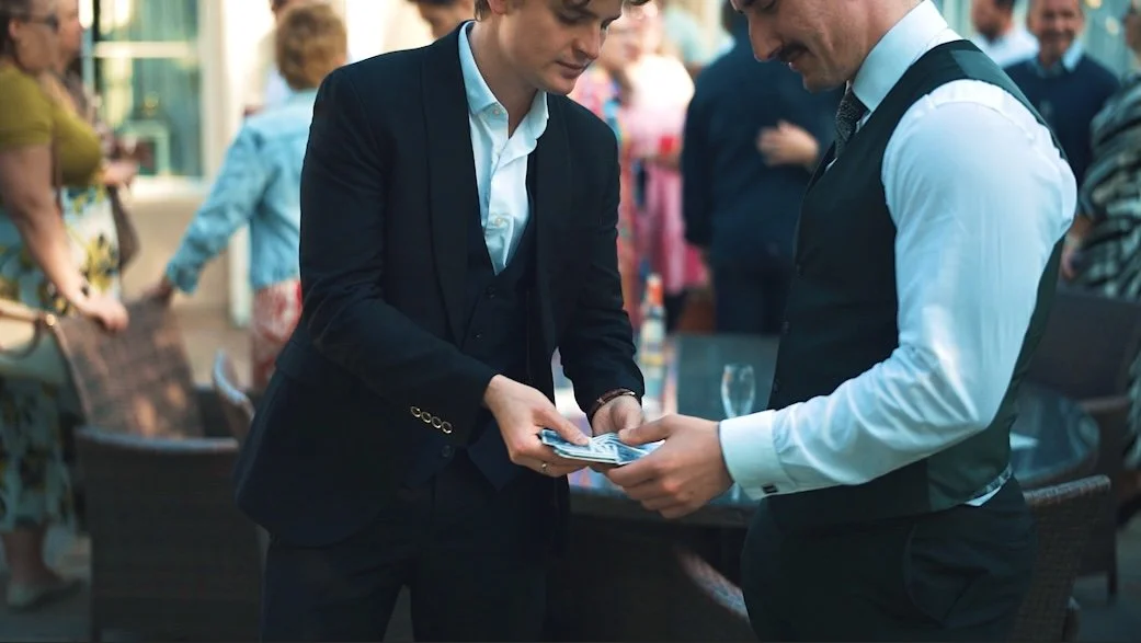 Two men in formal attire exchanging a stack of money at a social gathering or party.