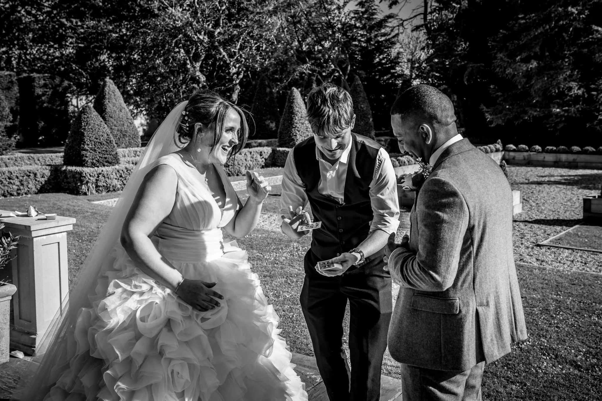 Black and white photo of a wedding scene with a bride, a groom, and  Daniel Maze performing a magic trick outdoors. The bride is smiling and pointing at the groom, who is looking at the magician. The magician is holding playing cards and is laughing.