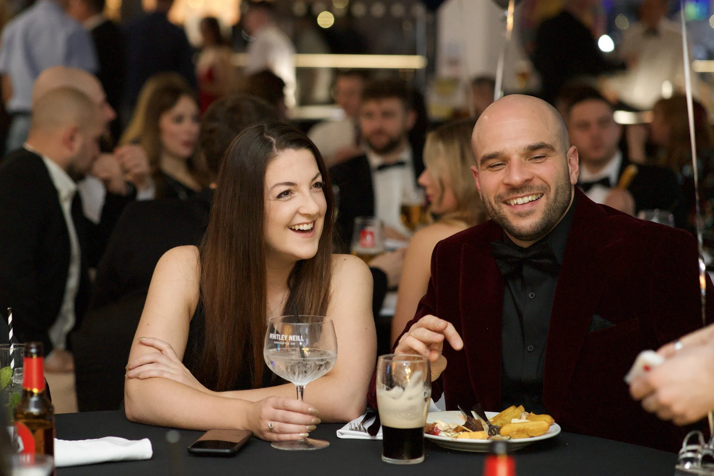 A man and woman smiling and sitting at a formal event with drinks and food on the table. Guests at Christmas do. 