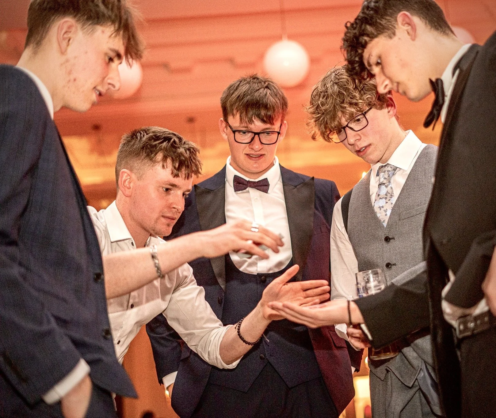 Group of well-dressed young men in suits focused on a hand gesture, performing or watching a magic trick, at an indoor Wedding. 