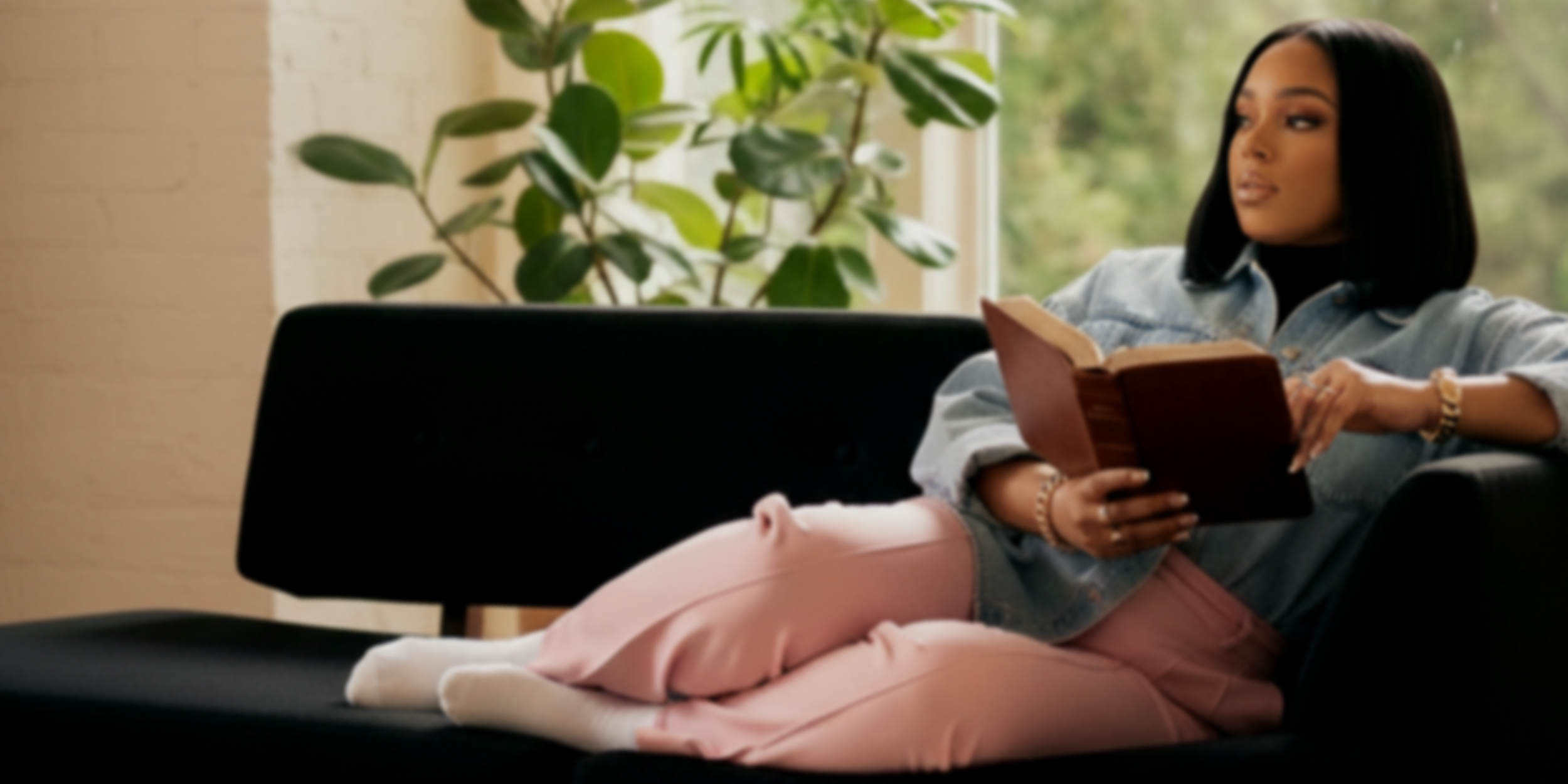 A woman with black hair, wearing a denim jacket and gold jewelry, laying on a dark sofa, reading a book, with a plant in the background near a window.