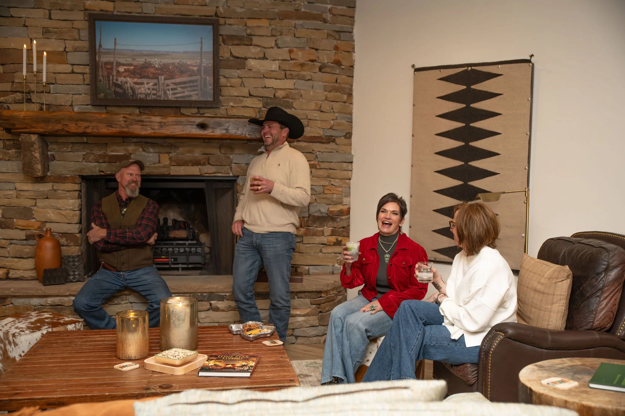 A group of people gathered in a cozy living room, with some standing and others sitting on a leather sofa. They are laughing and chatting, holding drinks. The background features a rustic stone fireplace, a wooden mantel, and wall art. A large painting of a landscape hangs above the mantel. The overall atmosphere is warm and cheerful.