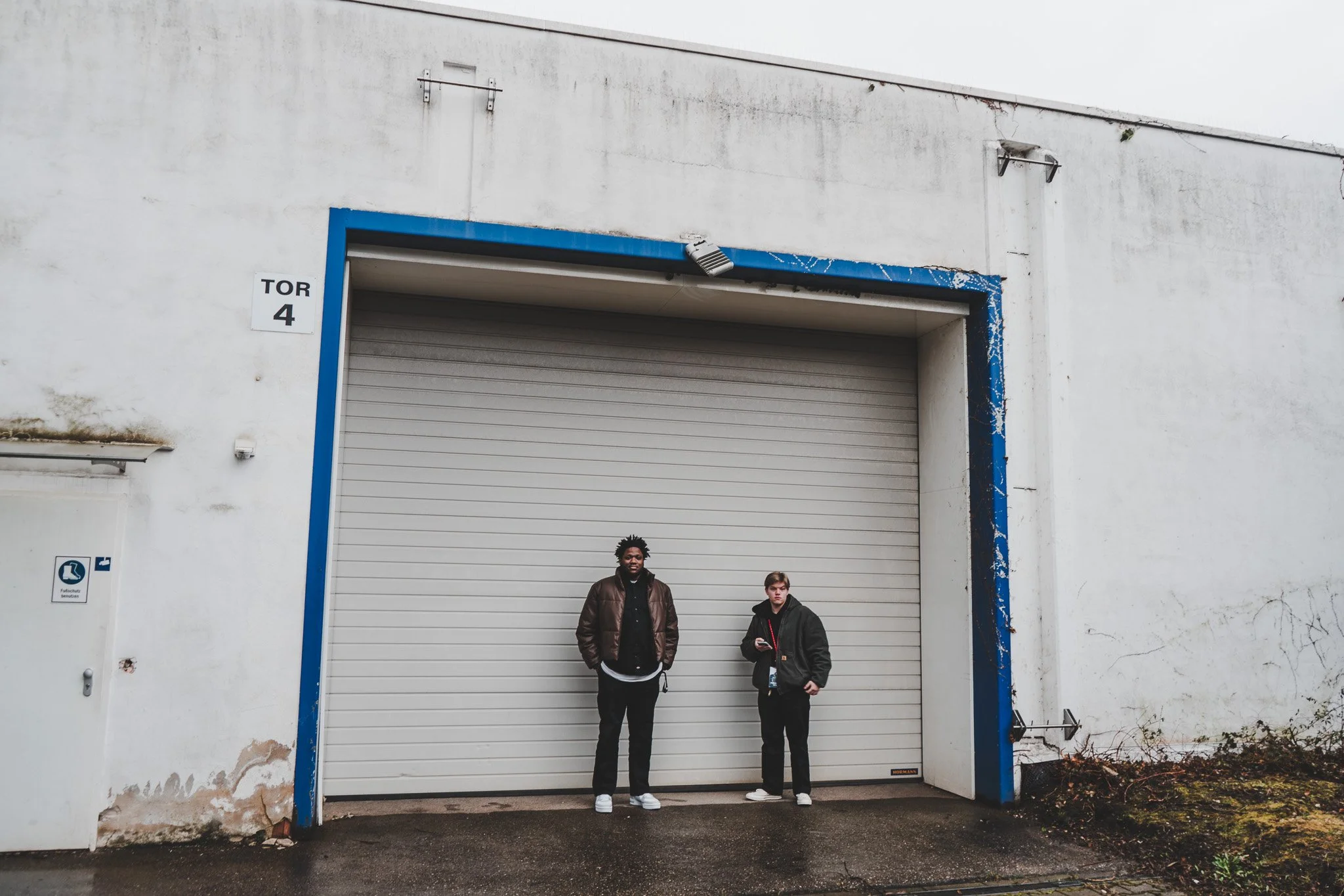Two young men standing in front of a large closed garage door with white walls, one in a brown jacket and the other in a black hoodie, on a damp outdoor pavement.