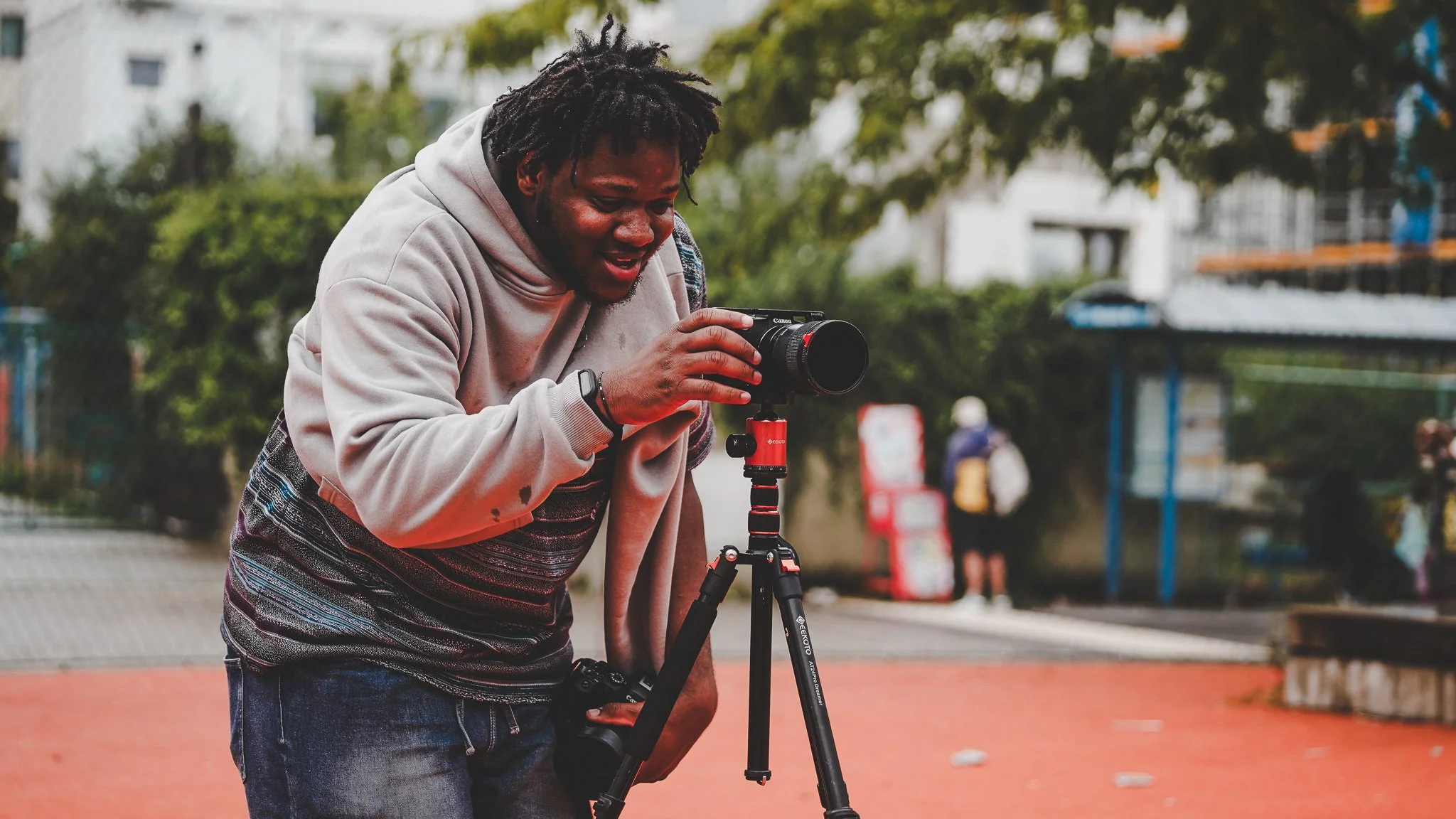 A man using a camera on a tripod outdoors in a park, with trees and a fence in the background.