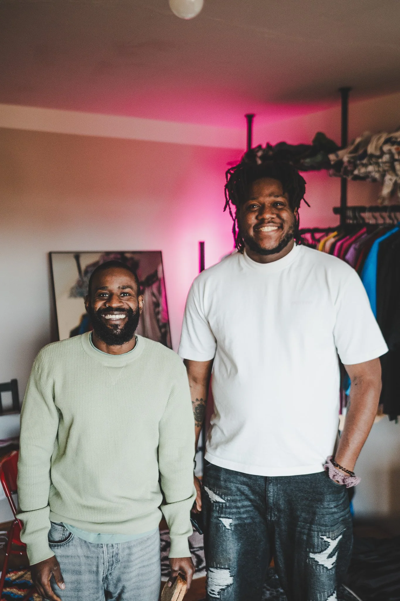 Two smiling men standing inside a room with clothing racks, a mirror, and pink lighting in the background.